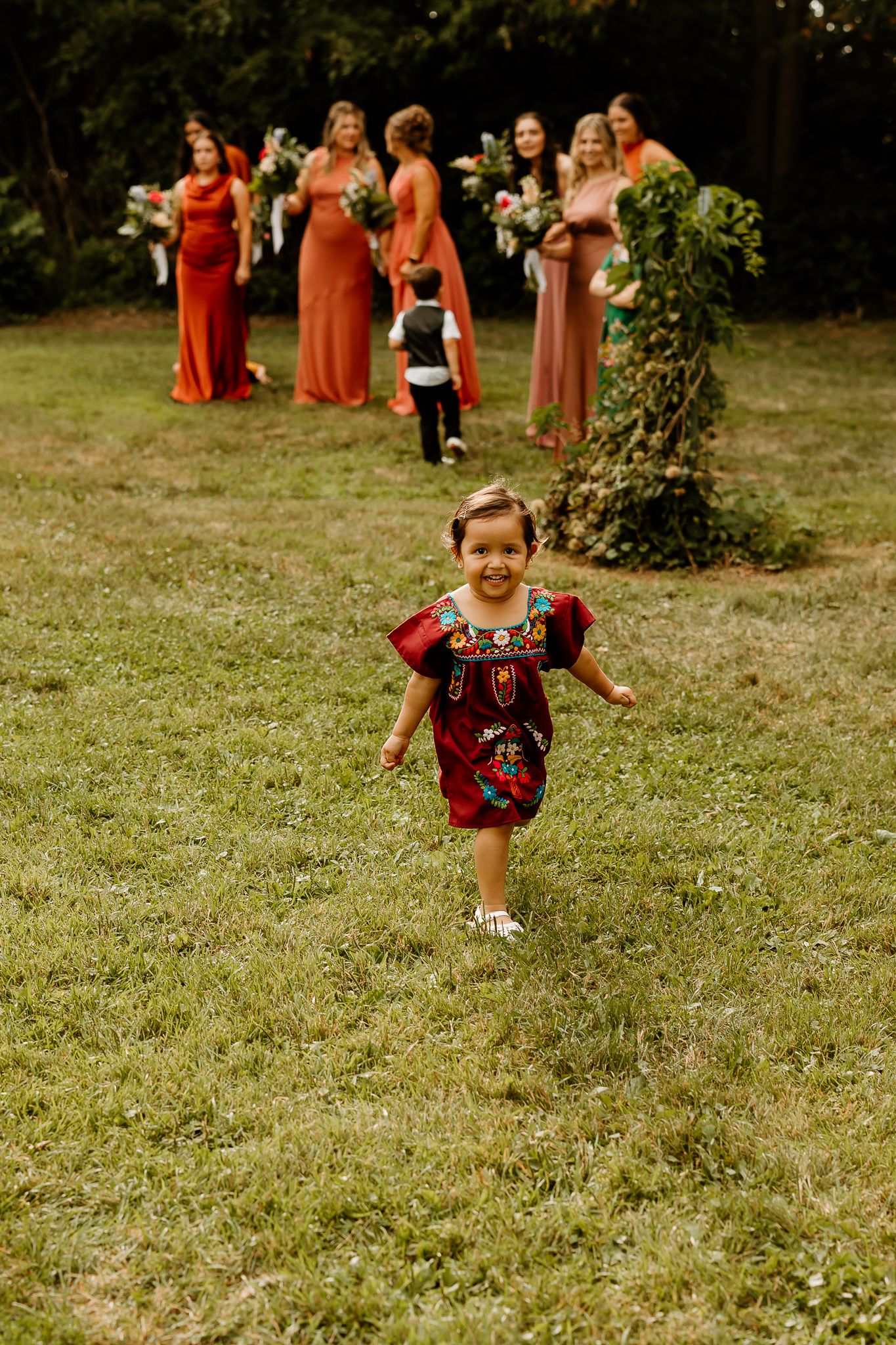 A joyful toddler in a red embroidered dress runs across green grass as bridesmaids in coral gowns stand in the background.
