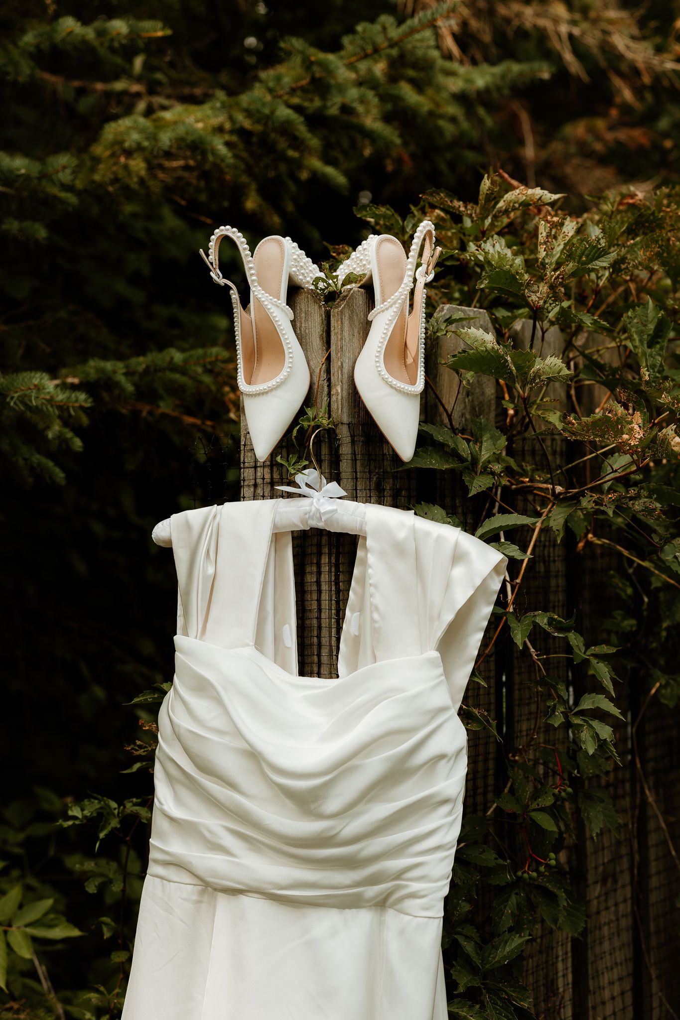 White wedding dress and pearl slingback heels displayed on a garden fence surrounded by lush green foliage.