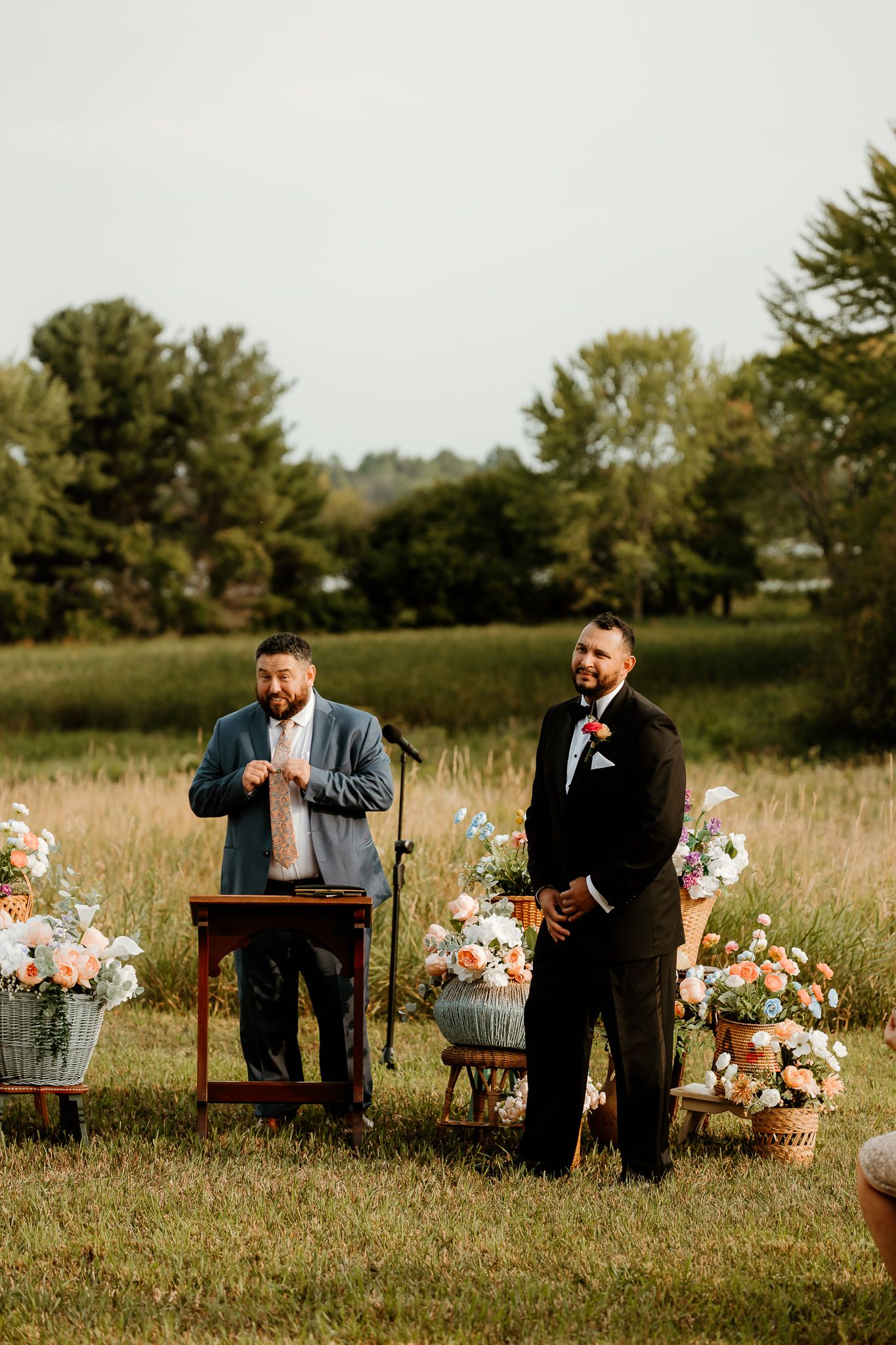 Groom in black tuxedo stands at outdoor wedding ceremony altar surrounded by floral arrangements in a lush green field.