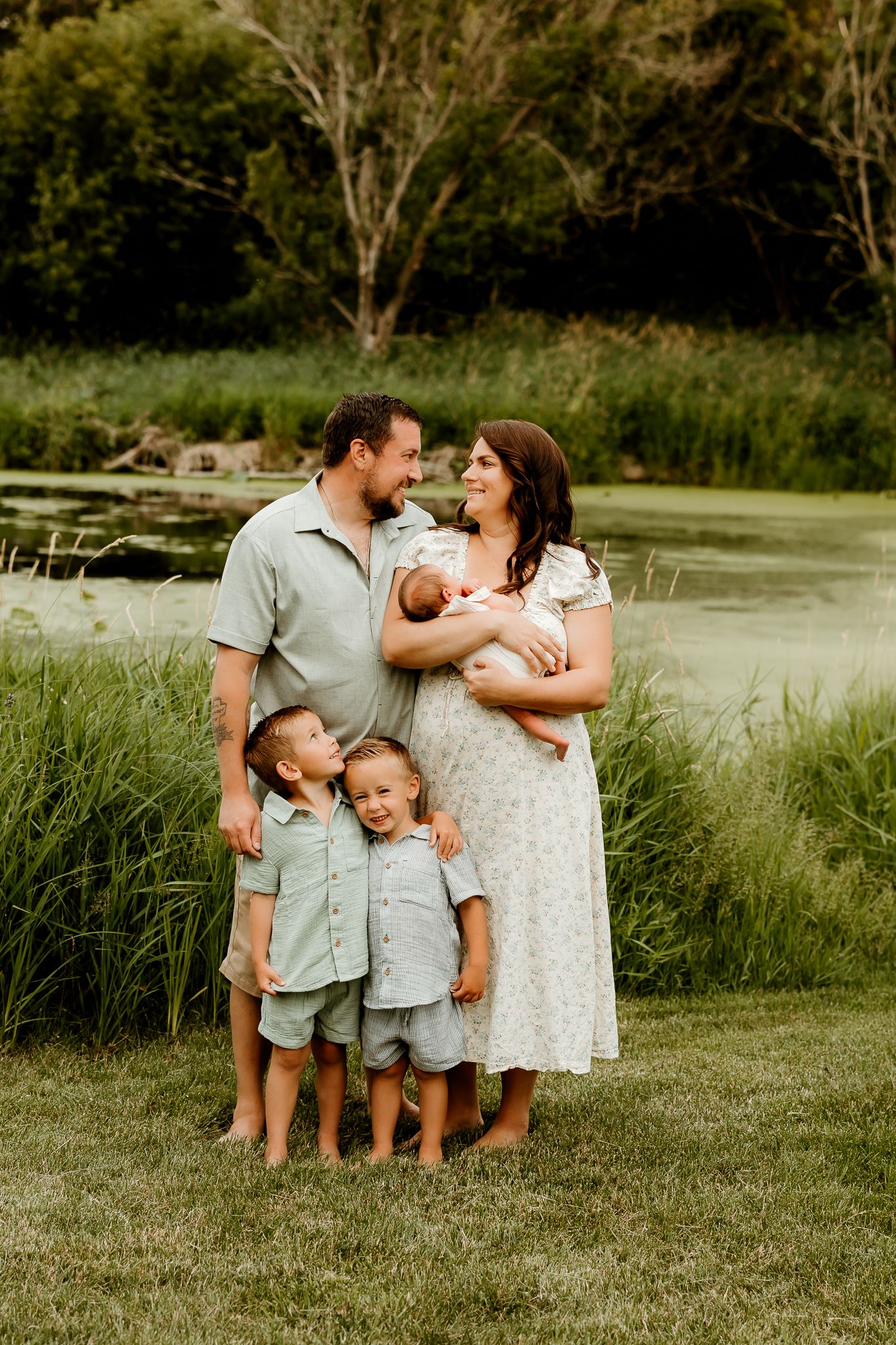 Happy family of five posing outdoors near a pond, parents with three young children including a newborn, in natural greenery.