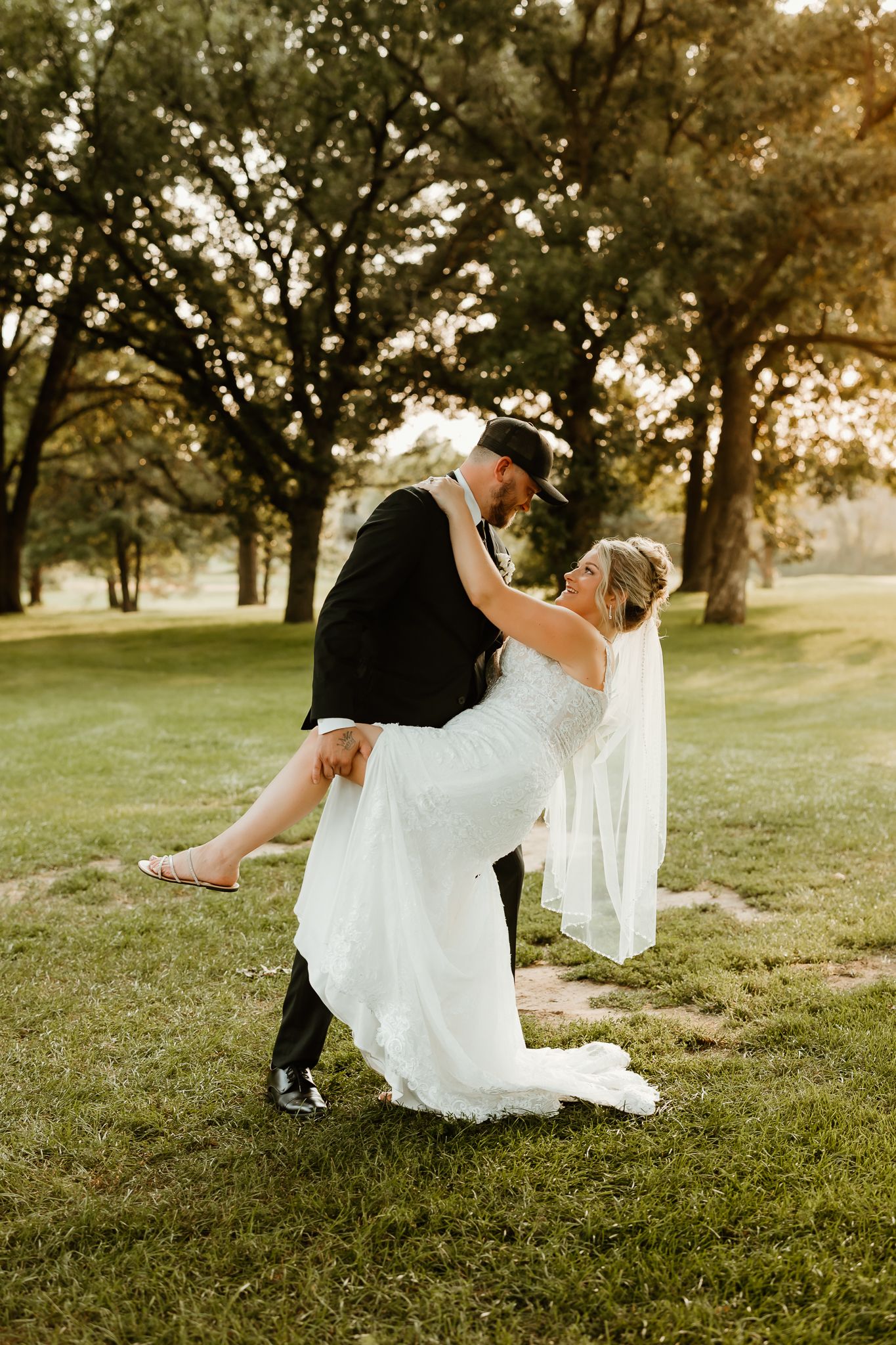 Groom lifting bride in white wedding dress outdoors on green grass with trees in golden hour light.