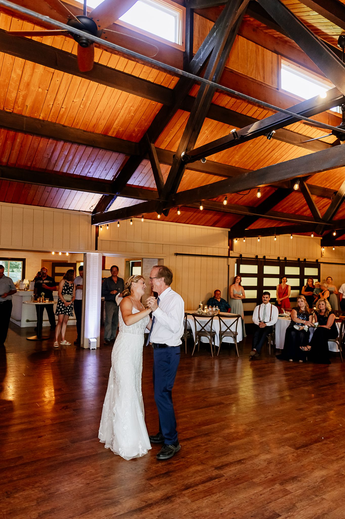 Bride and groom share first dance in rustic wood-beamed wedding venue with string lights and guests seated around dance floor.