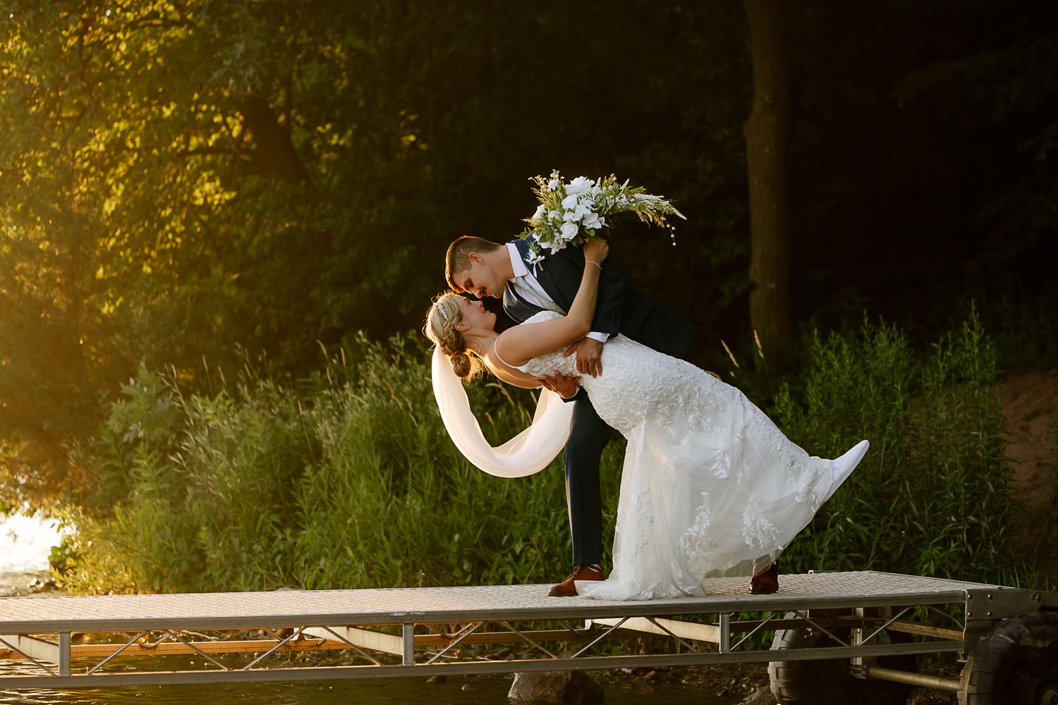 Bride and groom share a romantic dip kiss on a stage outdoors, surrounded by lush greenery in golden hour light.