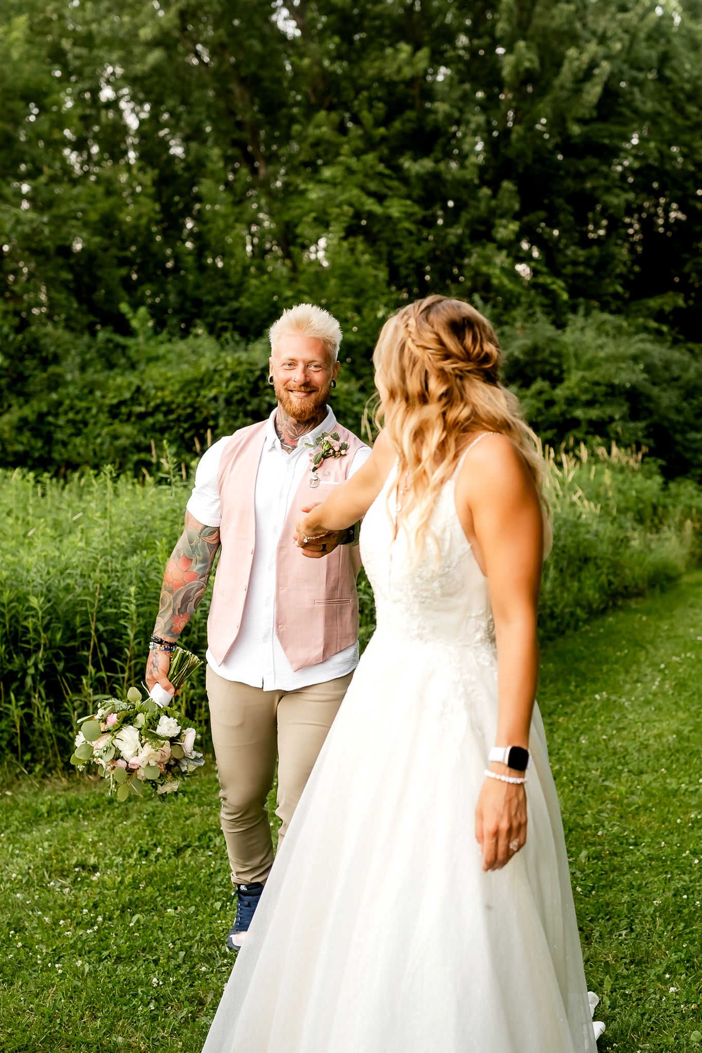 A tattooed groom in a pink vest holds his bride's hand outdoors on their wedding day amid lush greenery.