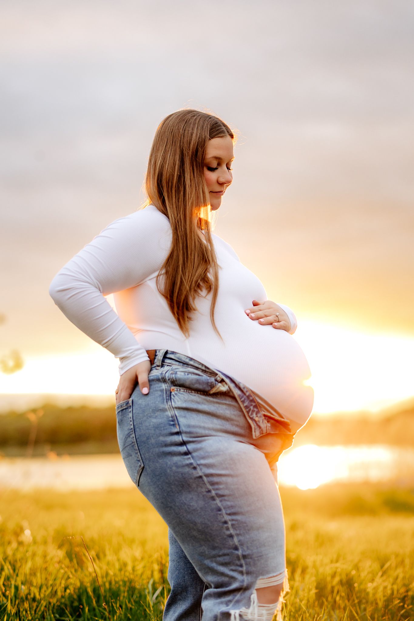 A pregnant woman in a white top and jeans cradles her baby bump during a golden sunset outdoors.