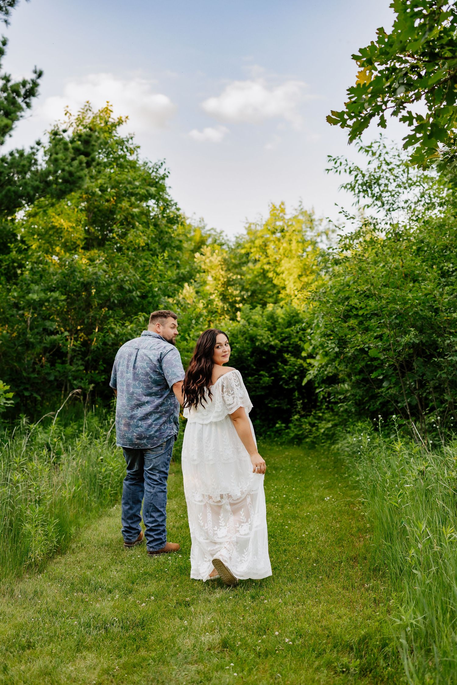 Couple holding hands walking through lush green meadow, woman in white lace maxi dress looking back at camera.