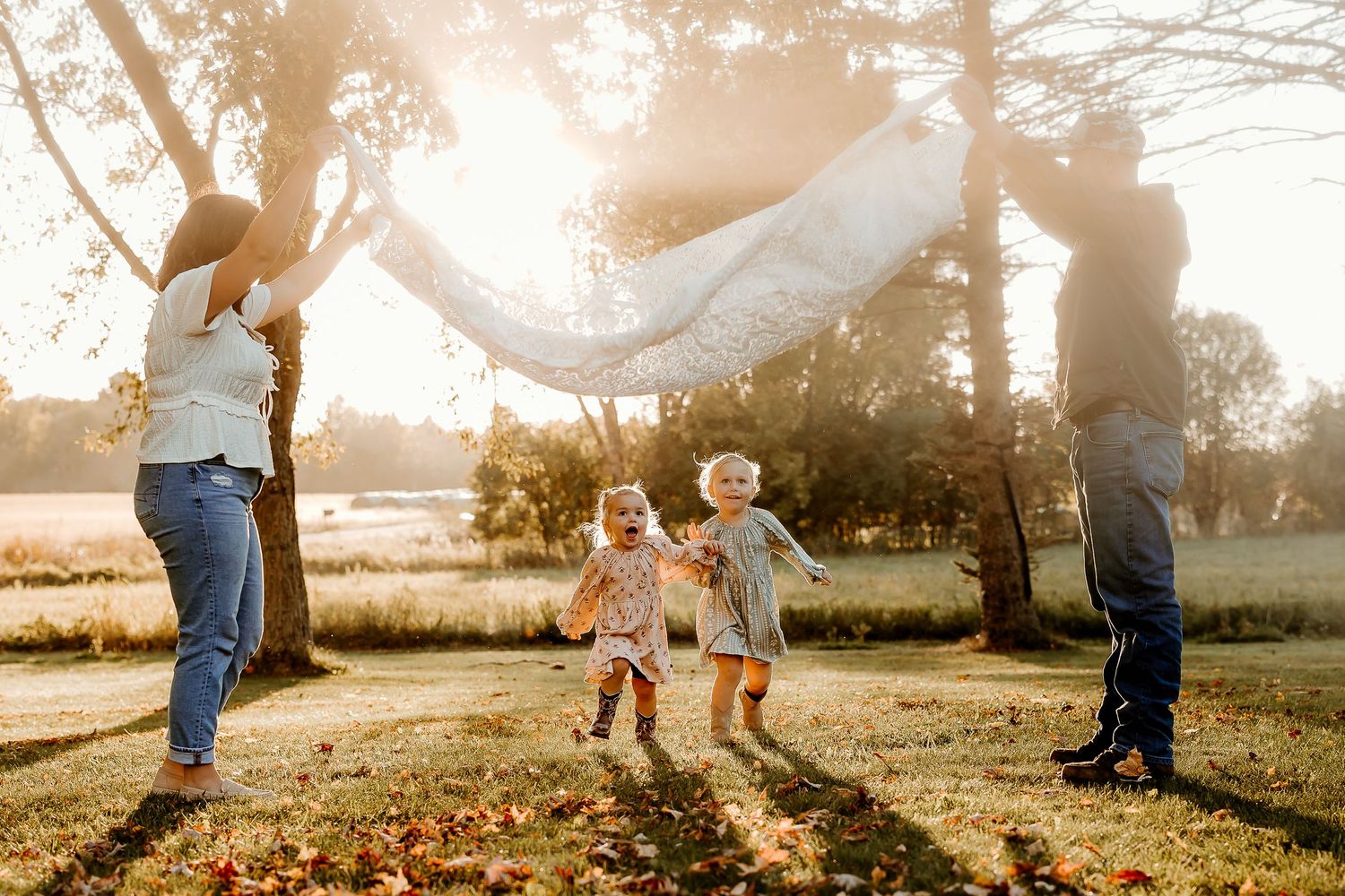 Family playing outdoors in golden sunlight, parents lifting a white blanket over two young children running on grass near trees.