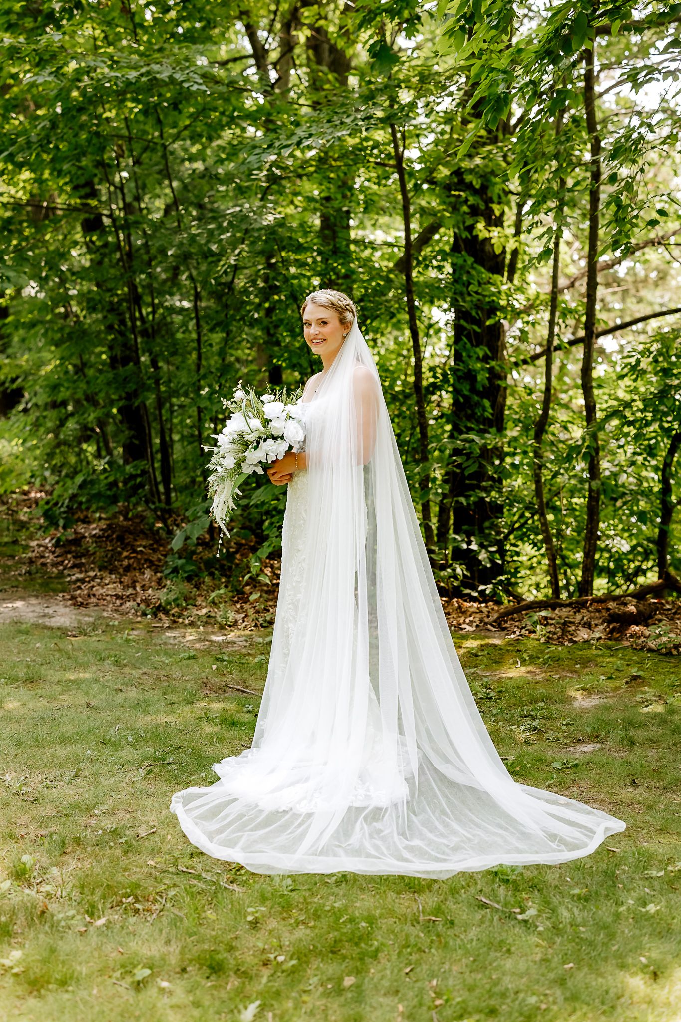 Bride in white gown and long cathedral veil holds bouquet outdoors among lush green trees.