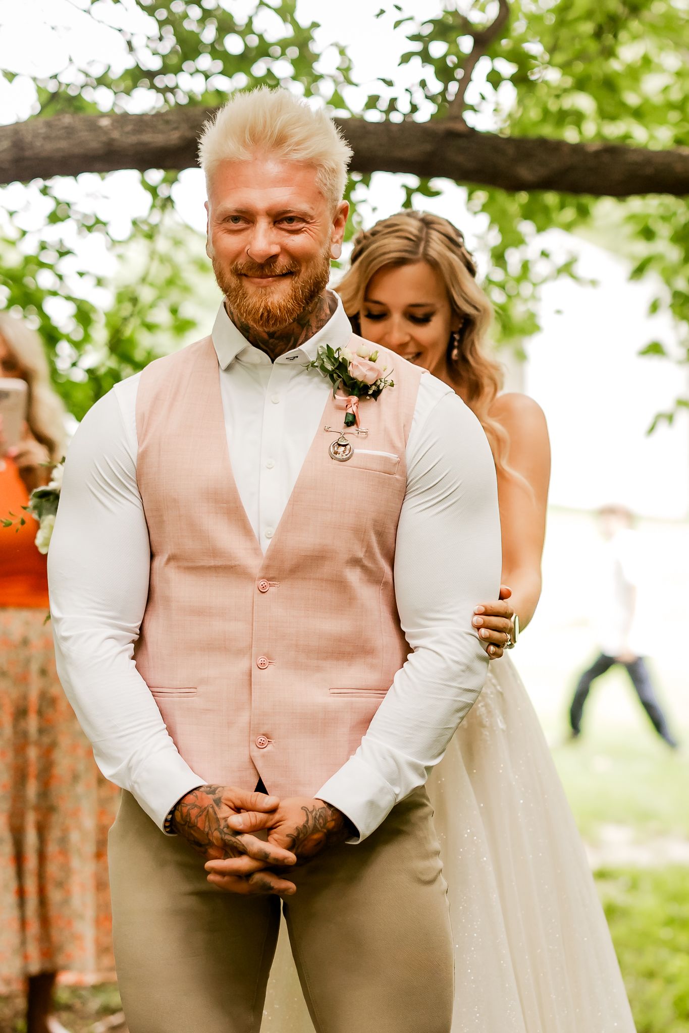 Smiling groom in pink vest with boutonniere stands with bride hugging him from behind at outdoor wedding.