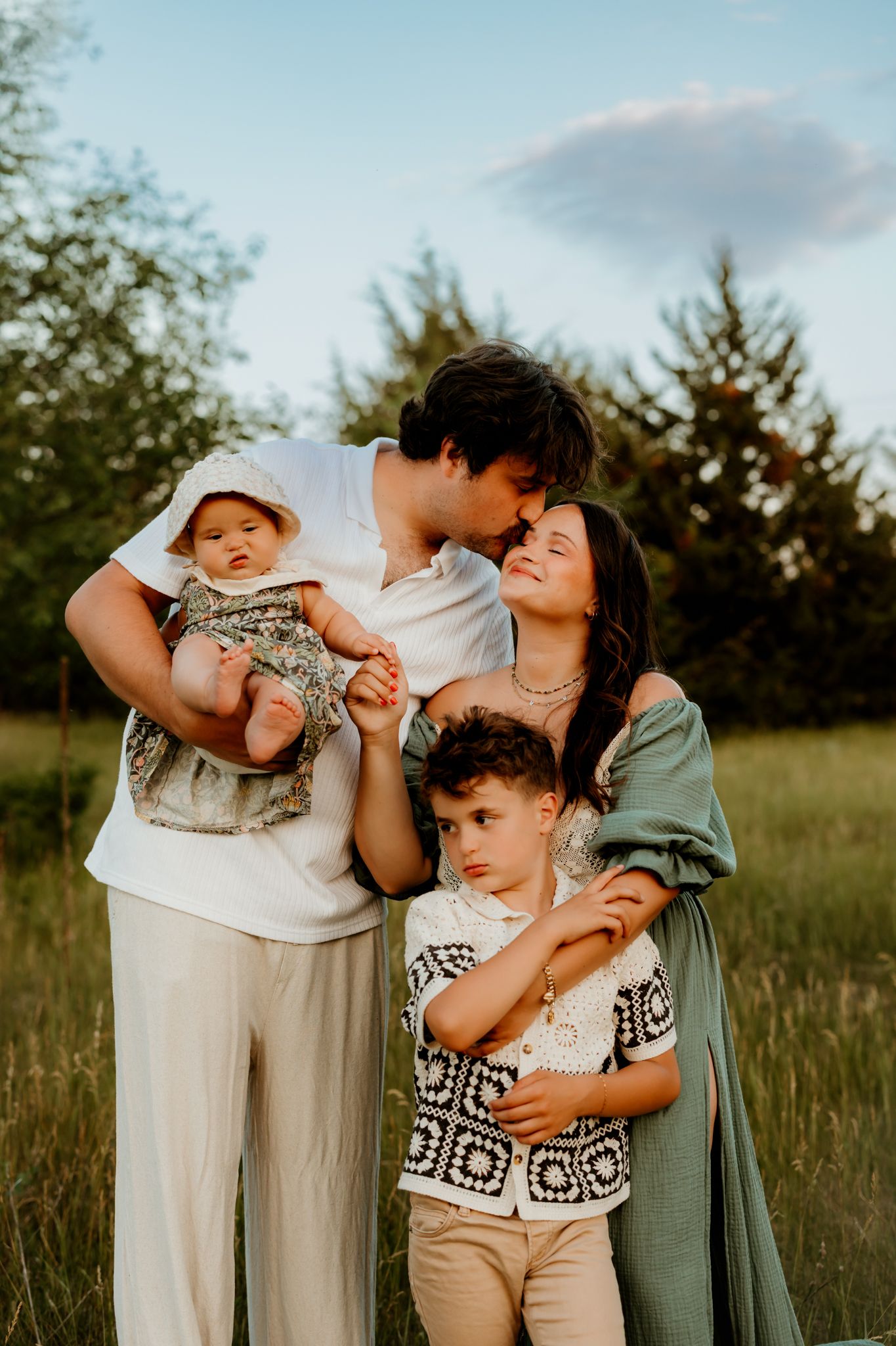 Family of four posing outdoors at golden hour, parents kissing while holding baby and young boy in green meadow.