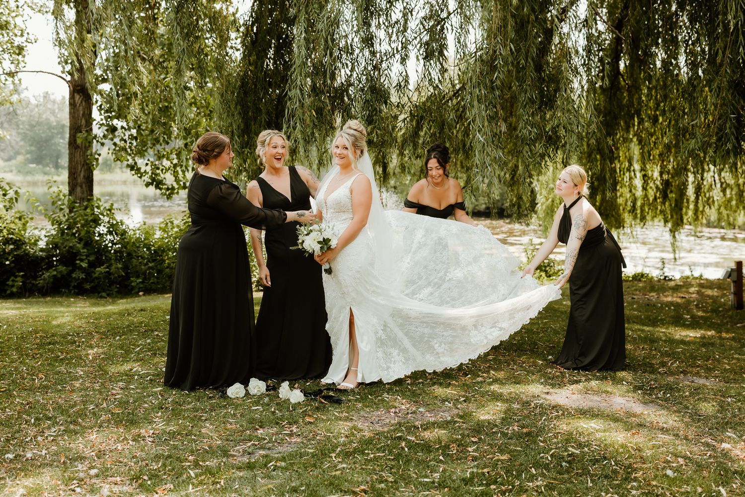 Bride in white gown with bridesmaids in black dresses spreading her train under a willow tree outdoors.