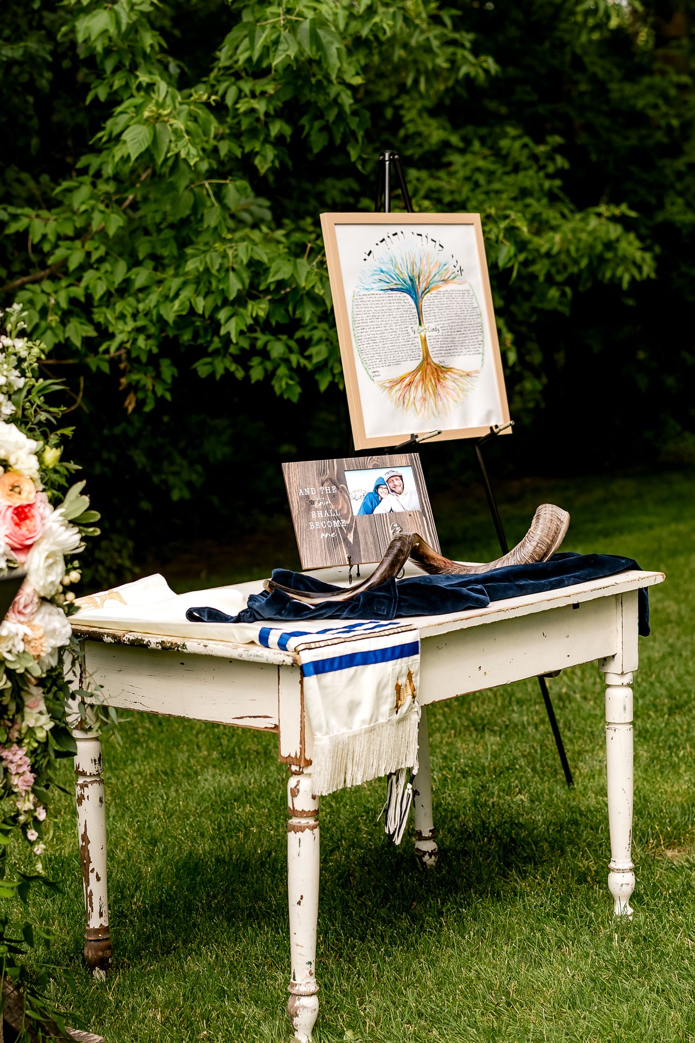 Rustic white vintage table with Jewish wedding items including a shofar, tallit, and tree of life artwork on easel outdoors.
