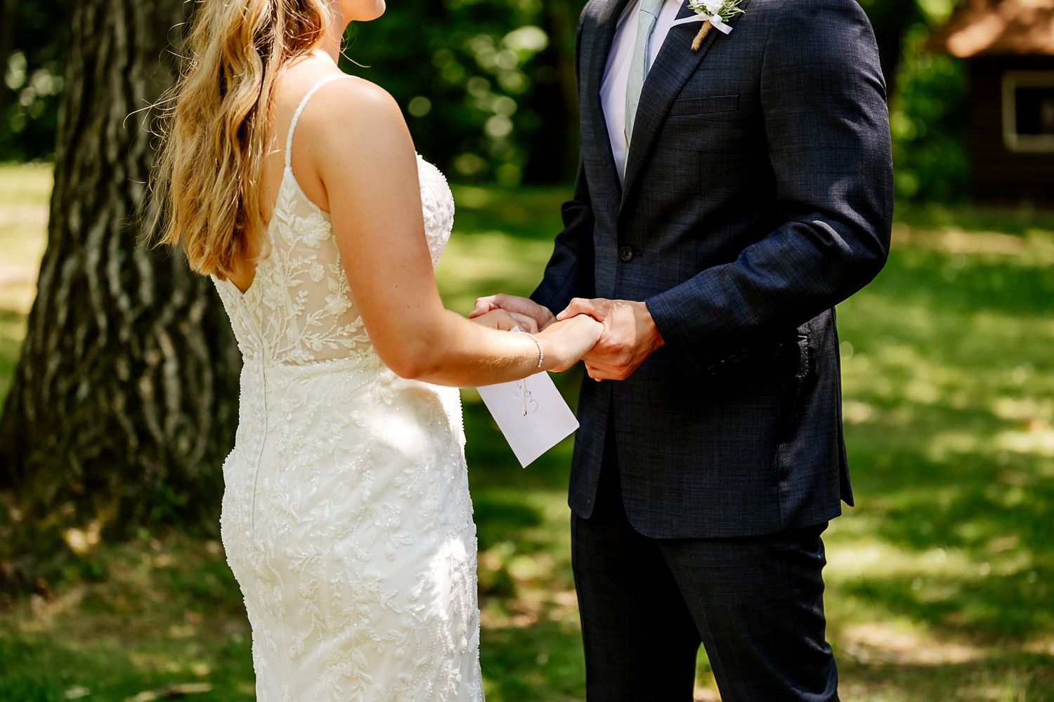 Bride and groom holding hands during outdoor wedding ceremony, bride in white lace gown, groom in navy suit.