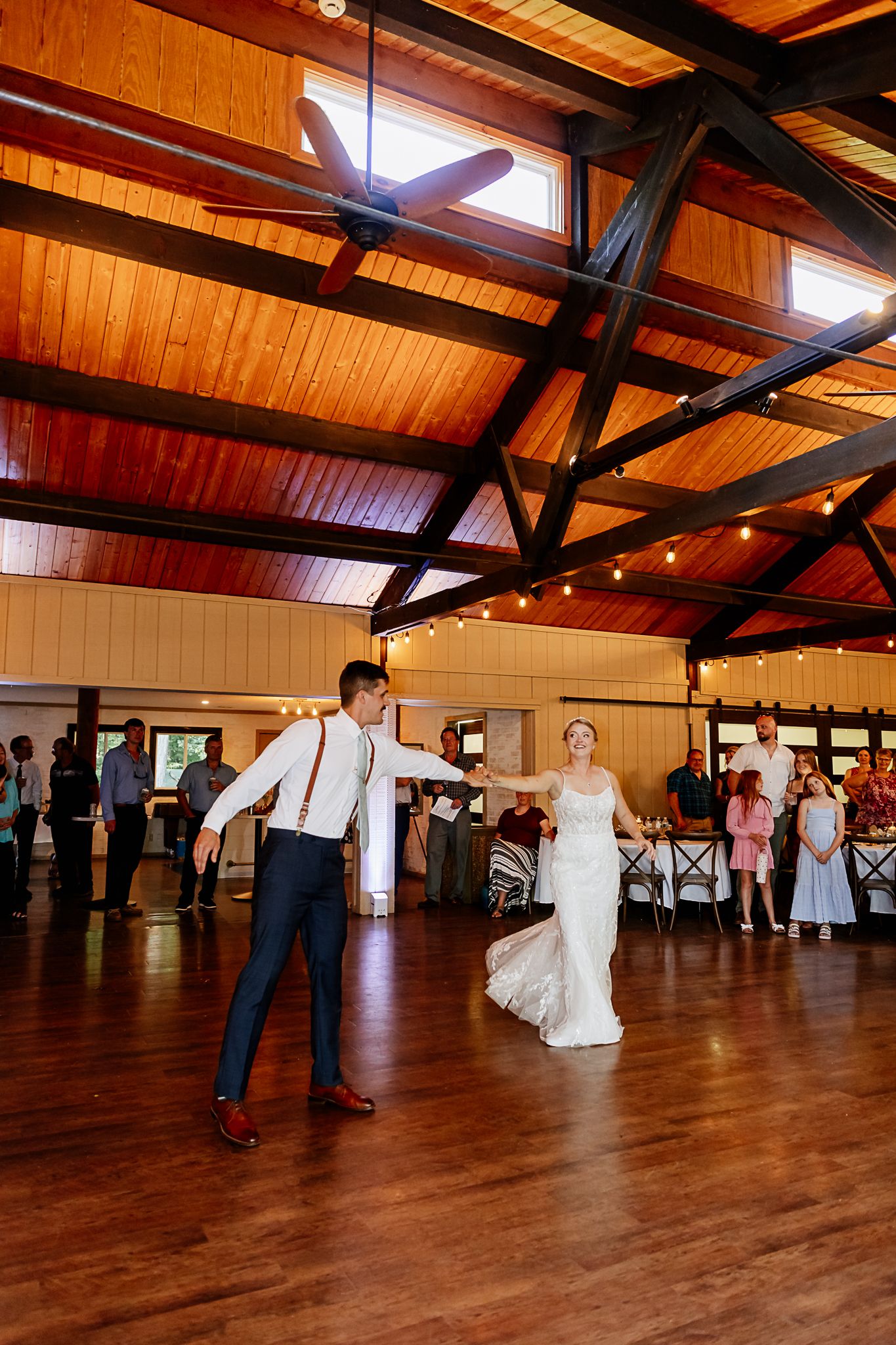 Bride and groom share their first dance on a wooden floor in a rustic venue with warm string lights.