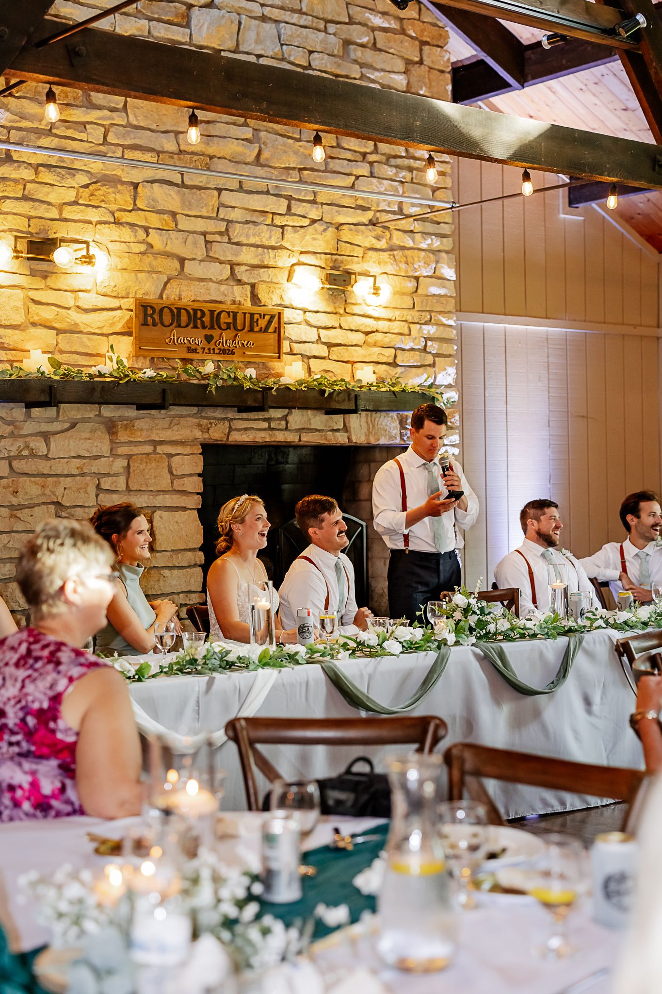 Wedding reception head table with bridal party, stone fireplace, Rodriguez sign, and elegant greenery decor in rustic venue.