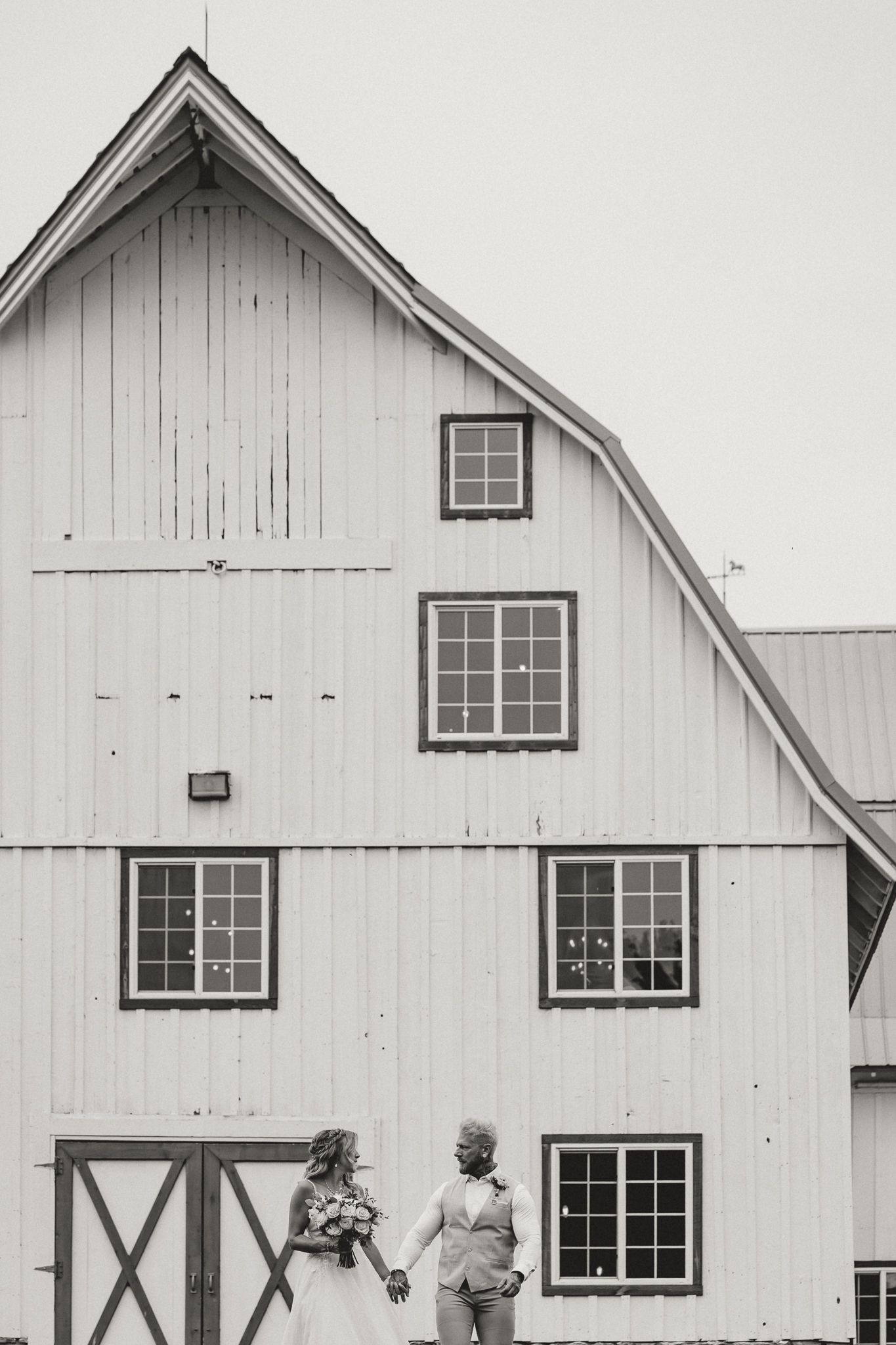 Black and white photo of a bride and groom holding hands in front of a large white barn wedding venue.