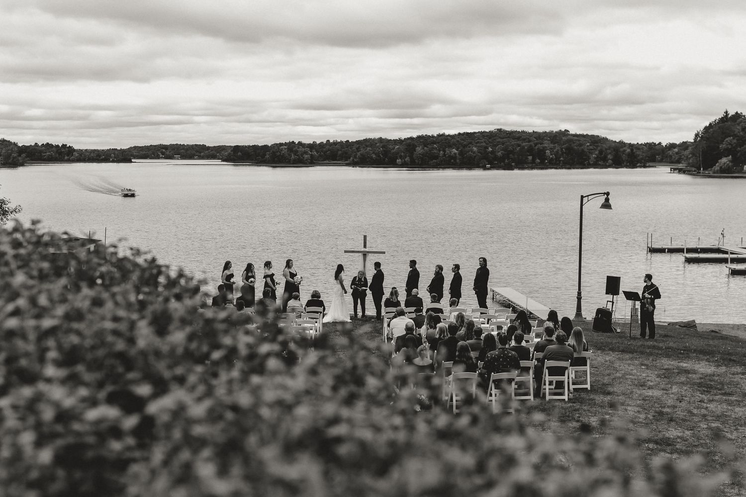 Black and white photo of an outdoor lakeside wedding ceremony with guests seated in rows facing the couple and officiant near the water.