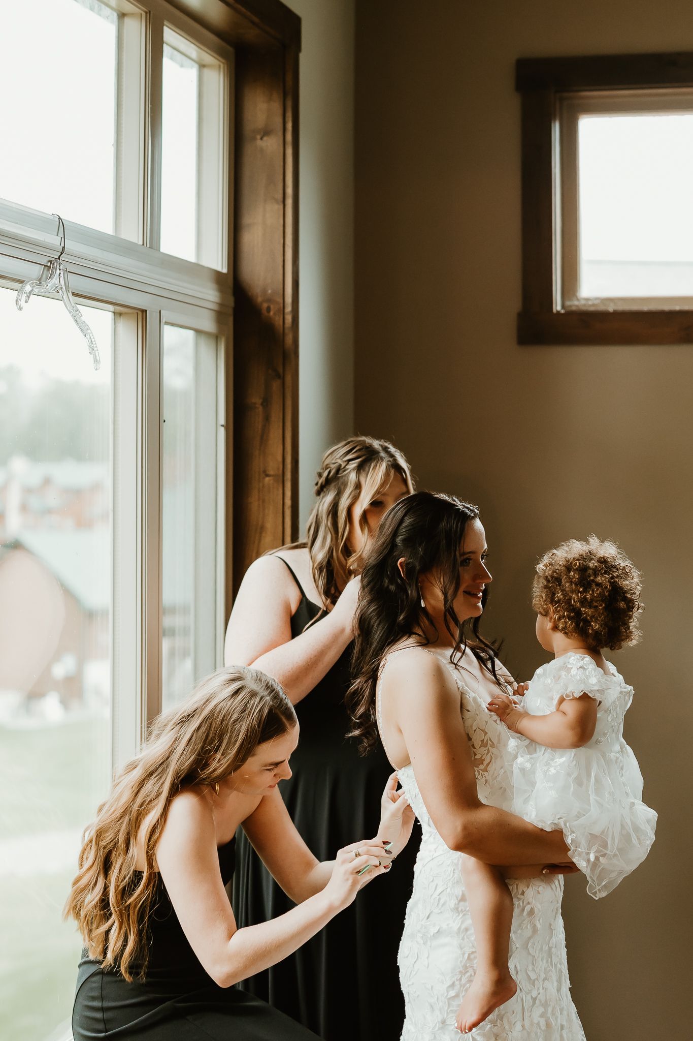 Two bridesmaids in black dresses help a bride in a lace wedding gown get ready by a sunlit window while she holds a toddler girl.