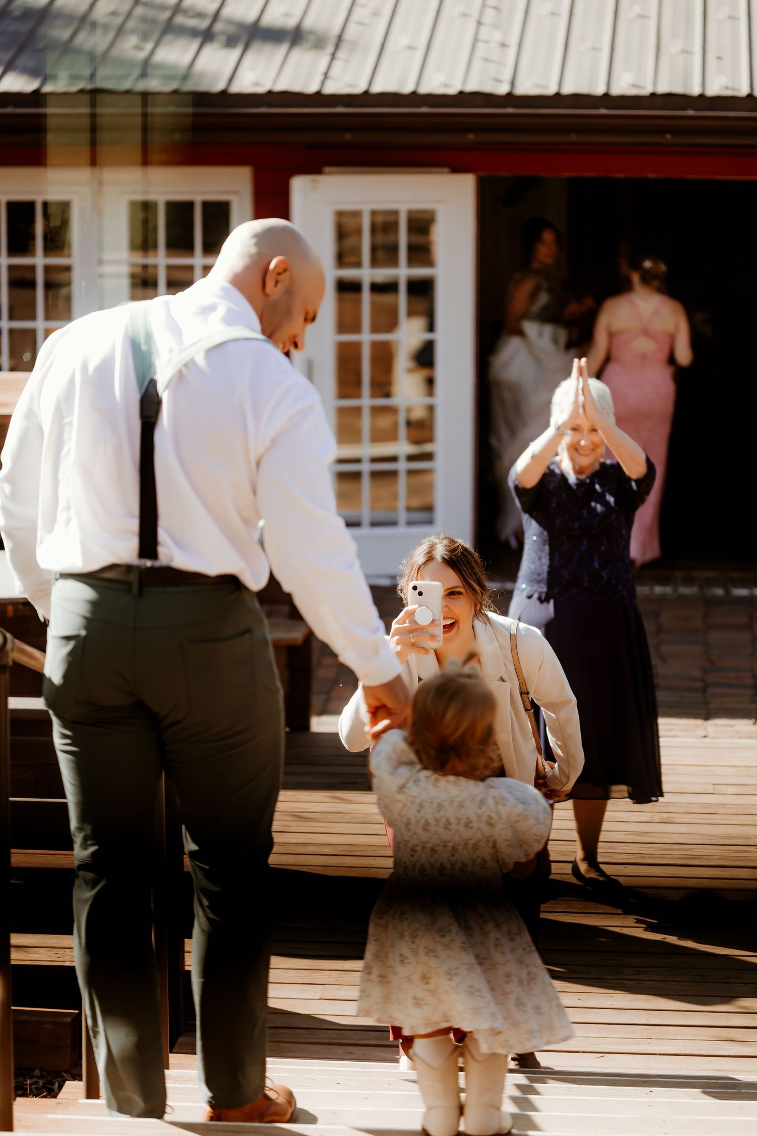 A man in suspenders holds hands with a toddler on a wooden deck as a smiling woman takes a photo and another woman claps nearby.