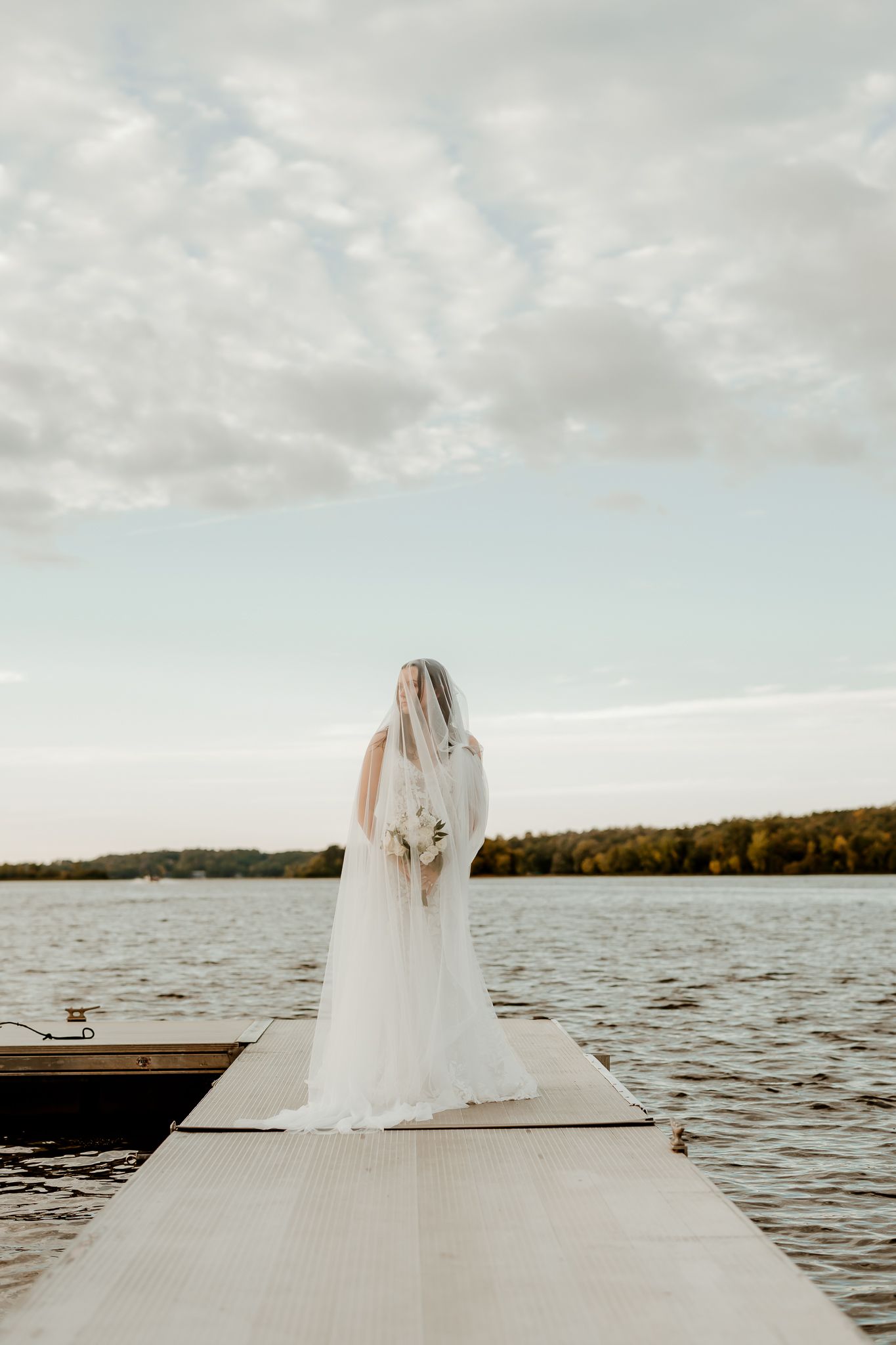 Bride in a long white gown and flowing veil holding a bouquet while standing on a wooden lake dock under a soft cloudy sky.