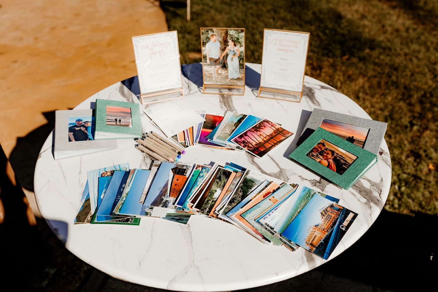 Round white table outdoors displaying scattered photo prints, framed pictures, and small signs at a wedding or event photo display.