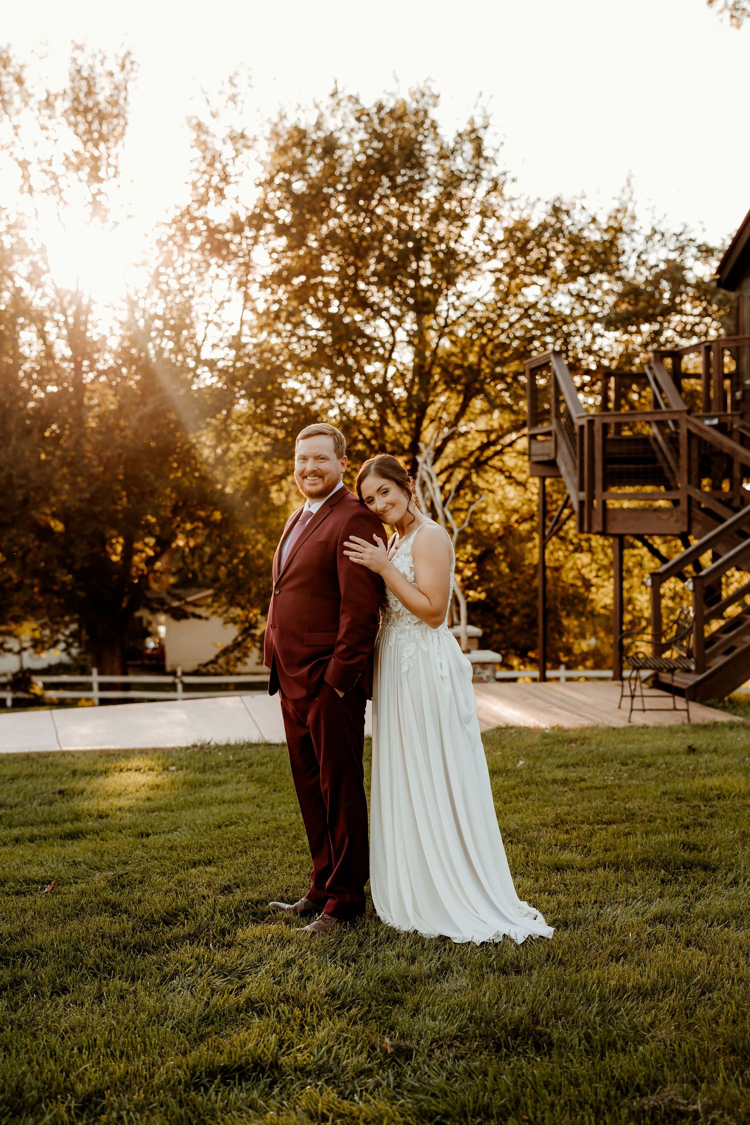 Smiling groom in a burgundy suit stands with his bride in a flowing white gown on a sunlit lawn near a wooden staircase at golden hour.