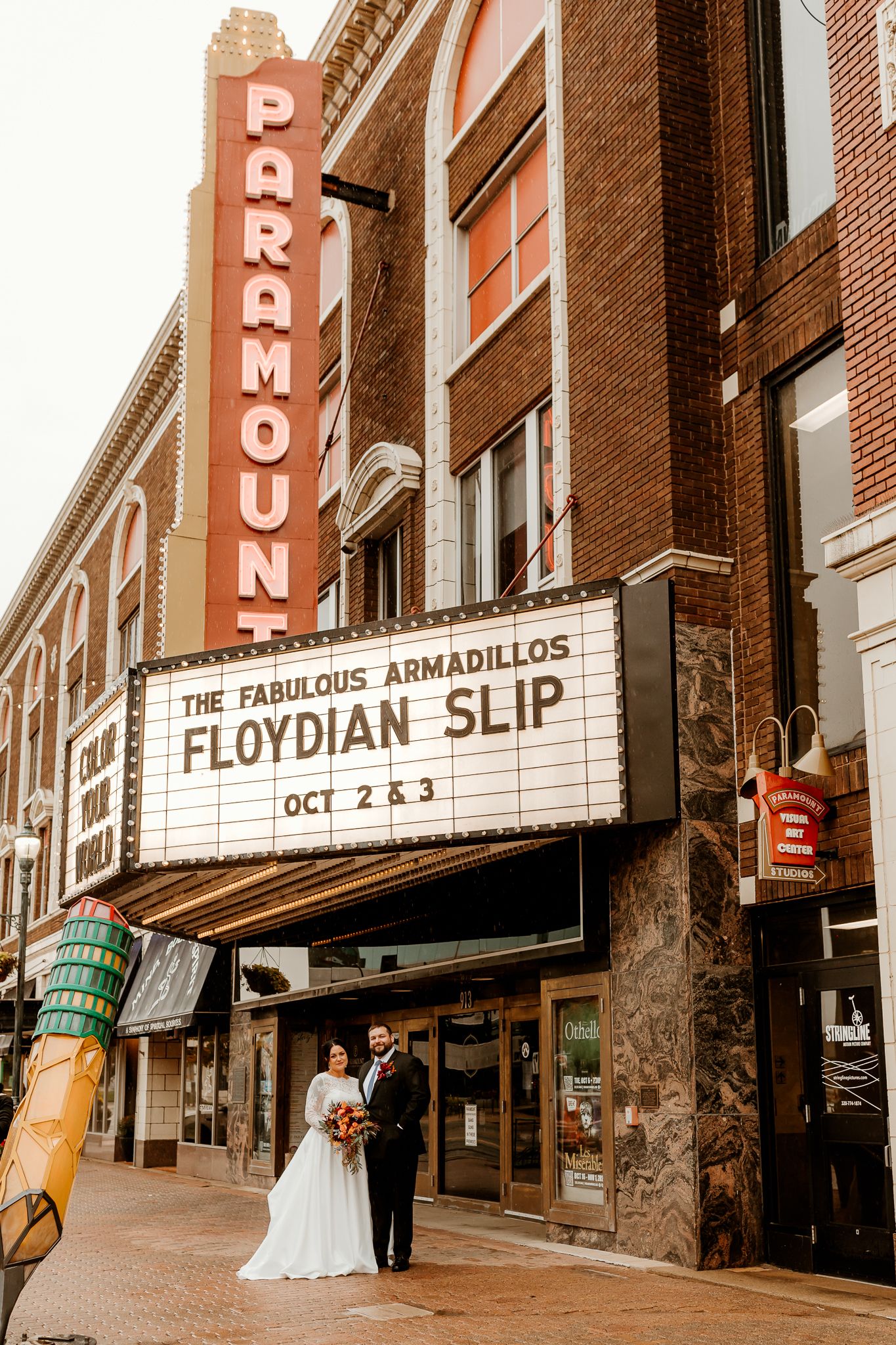 Bride and groom pose beneath the Paramount Theatre marquee advertising The Fabulous Armadillos and Floydian Slip on a brick-lined street.