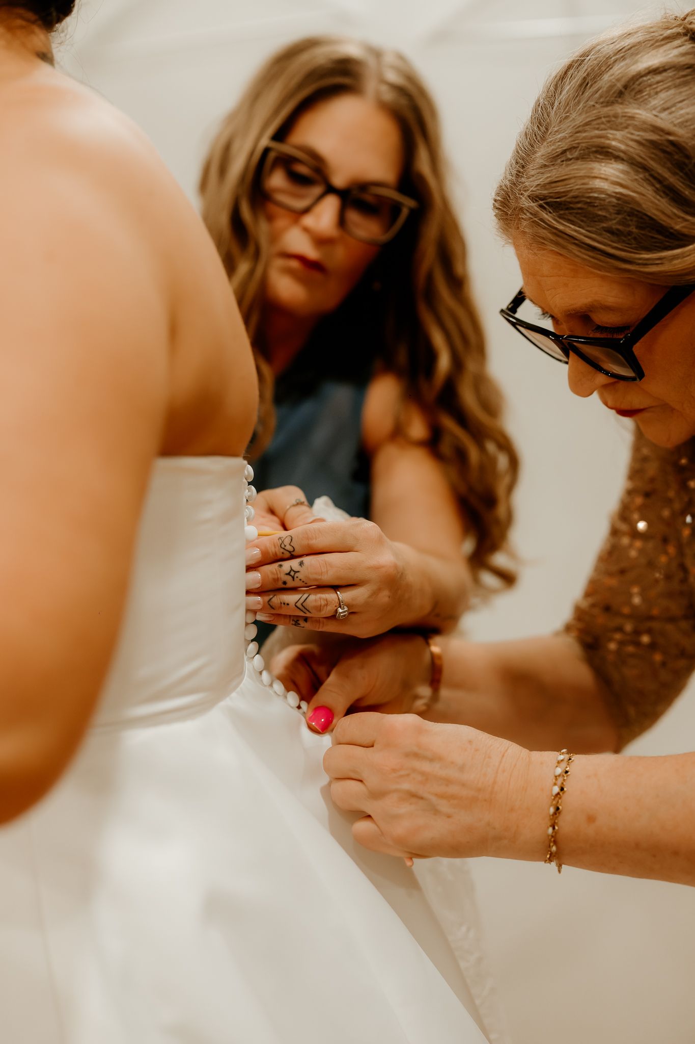 Two women with glasses help a bride button the back of her strapless white wedding gown during preparations before the ceremony.