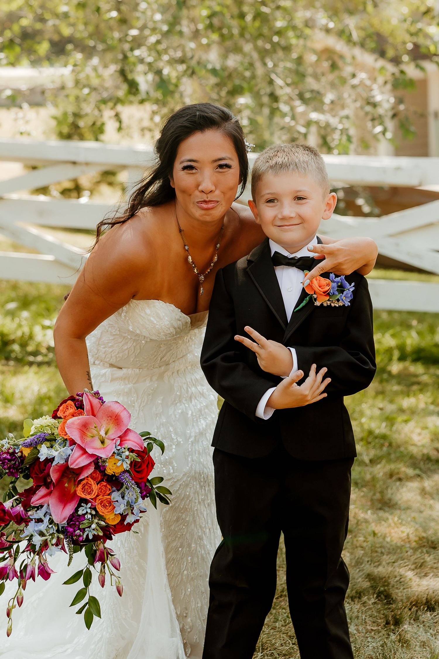 Bride in white gown holding colorful bouquet poses with young ring bearer in tuxedo at outdoor wedding.