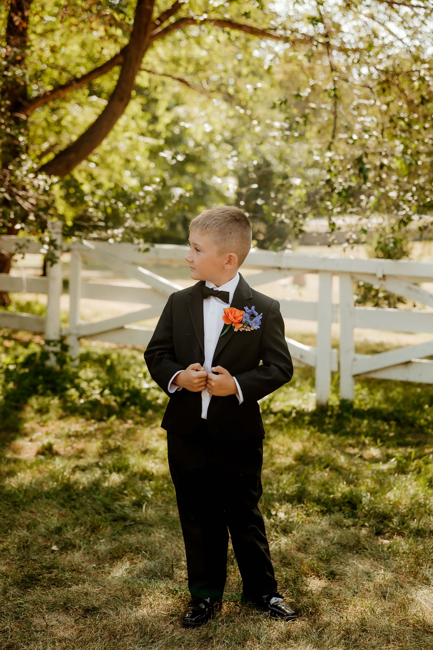 Young boy in black tuxedo with floral boutonniere stands outdoors near white fence at a wedding.