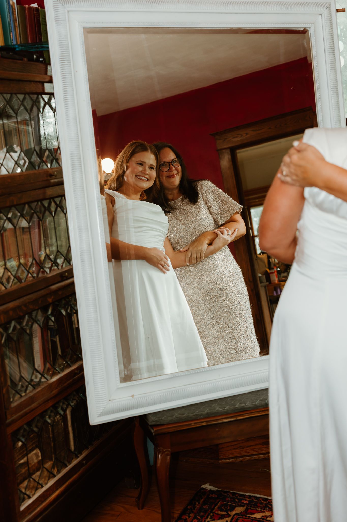 Bride in white wedding dress smiling with helper in mirror reflection inside elegant room with bookcase.