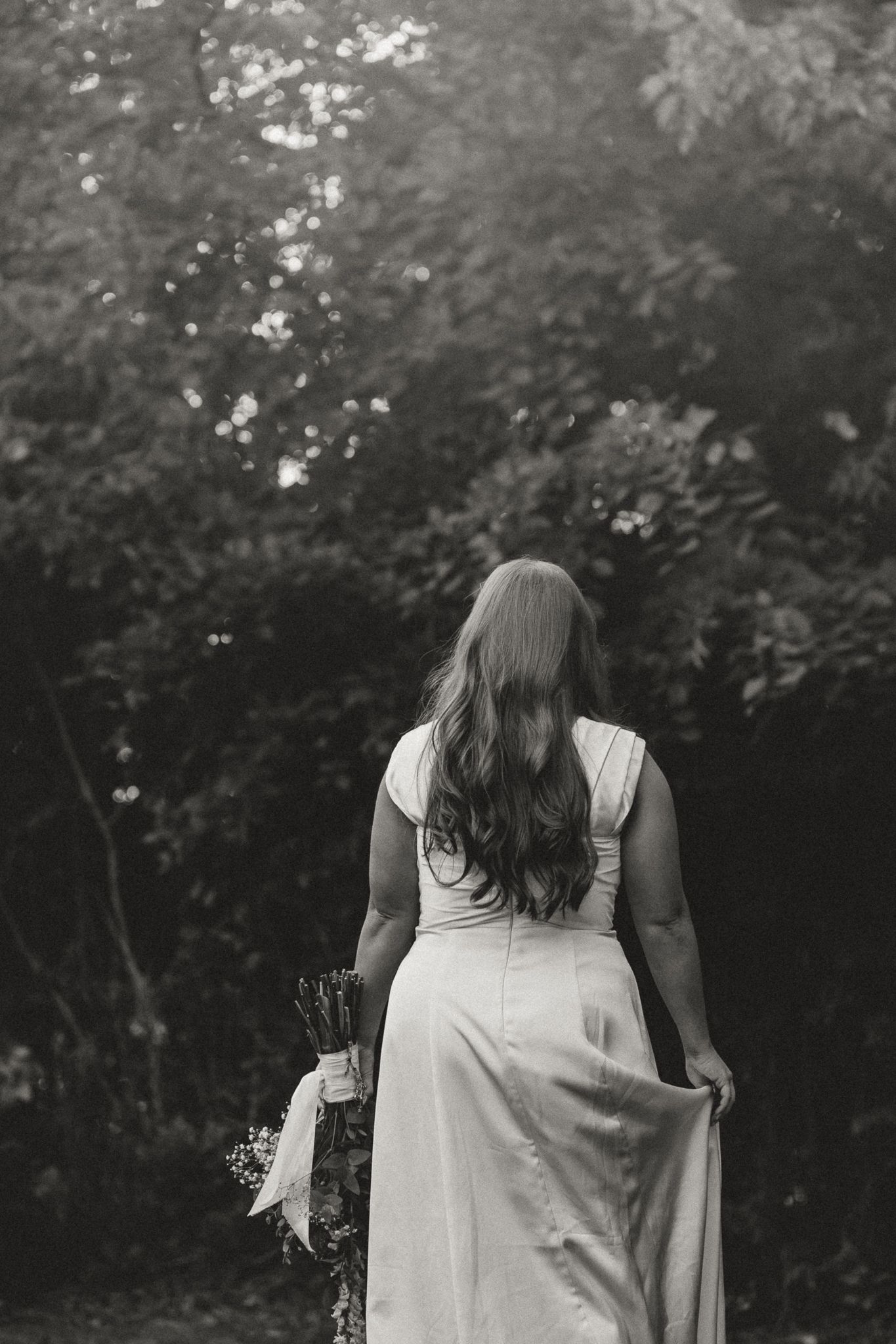 Black and white photo of a bride in a white gown holding a bouquet, standing with her back to the camera in a garden.