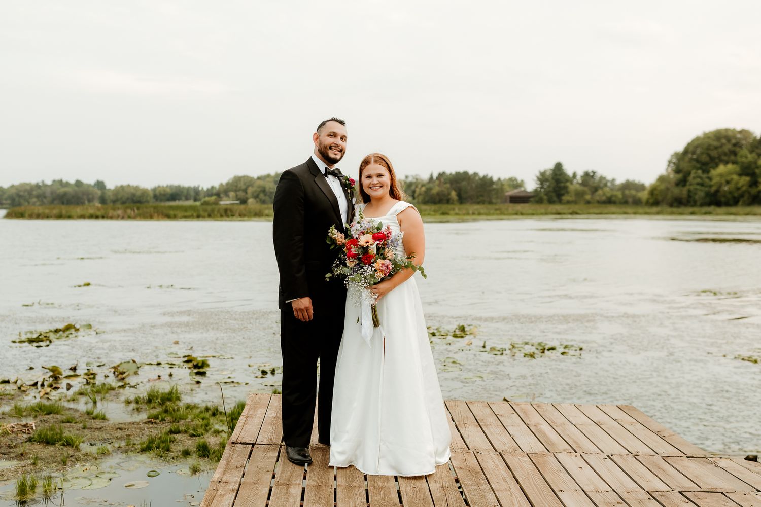 Bride and groom pose on a wooden dock by a serene lake, bride holding a red bouquet in a white gown.