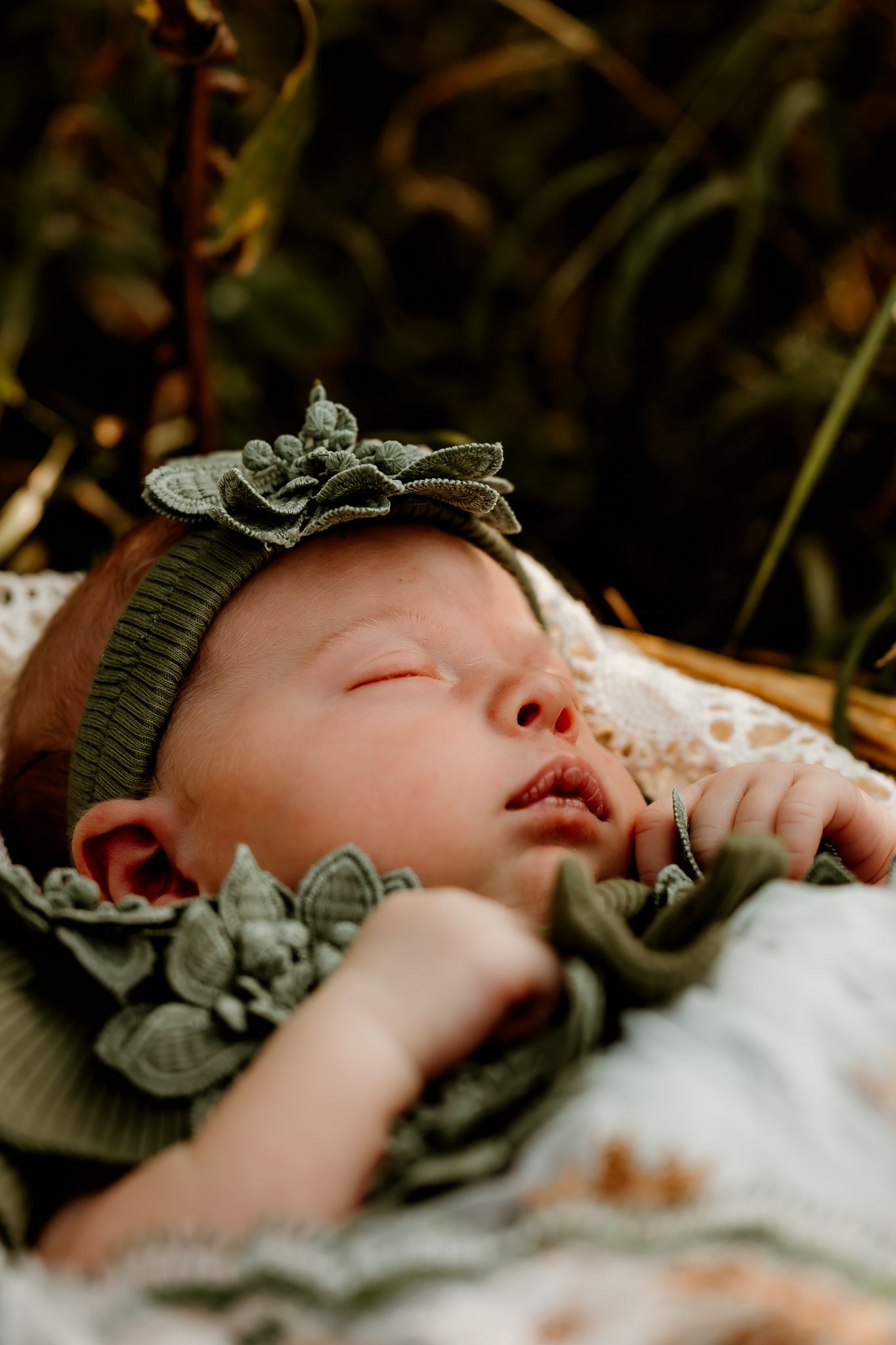 Sleeping newborn baby wearing a green floral headband and matching outfit, nestled in a basket outdoors.