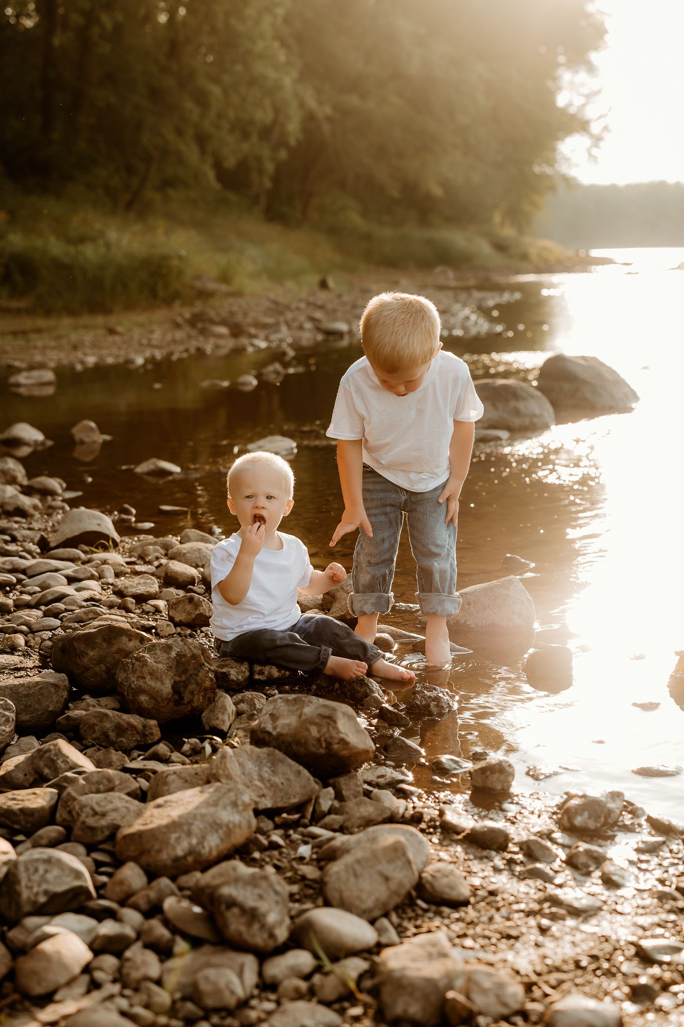 Two young boys playing on a rocky riverbank at golden hour, with trees and calm water in the background.