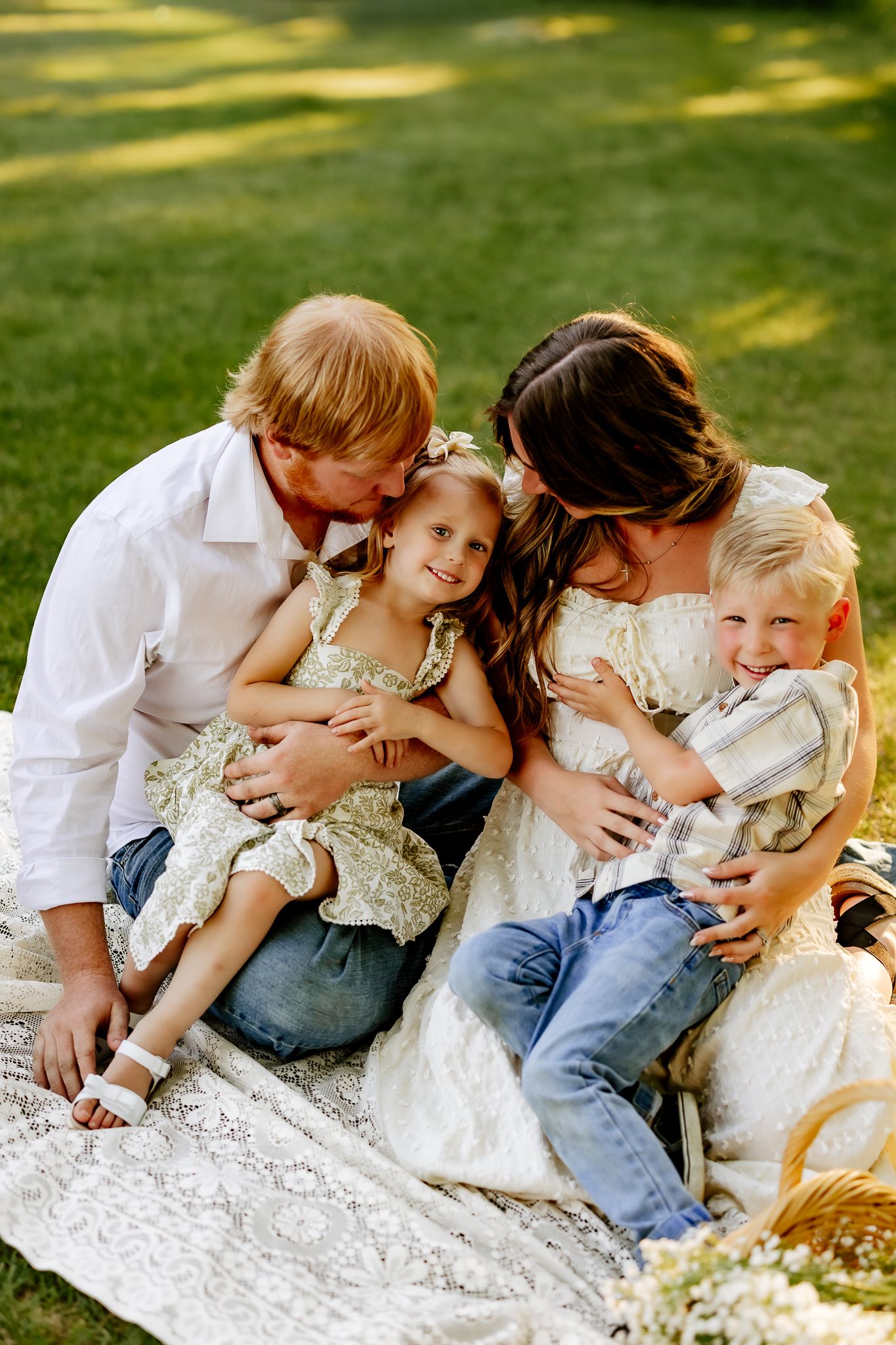 A joyful family of four sits on a lace blanket in green grass, parents kissing their two young children.