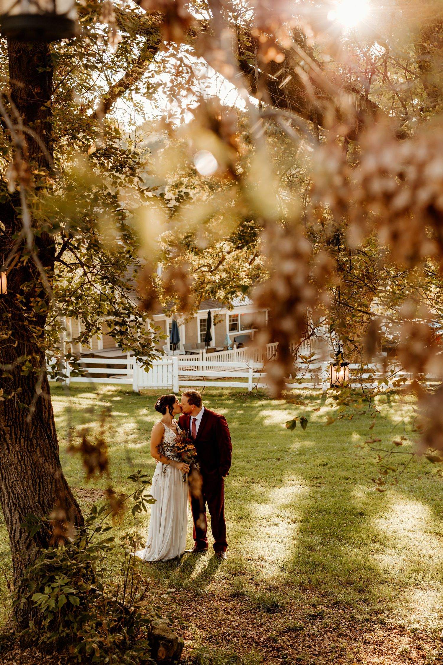 Bride in a white gown and groom in a dark suit kiss on a sunlit lawn framed by tree branches, with a white fence and house behind.