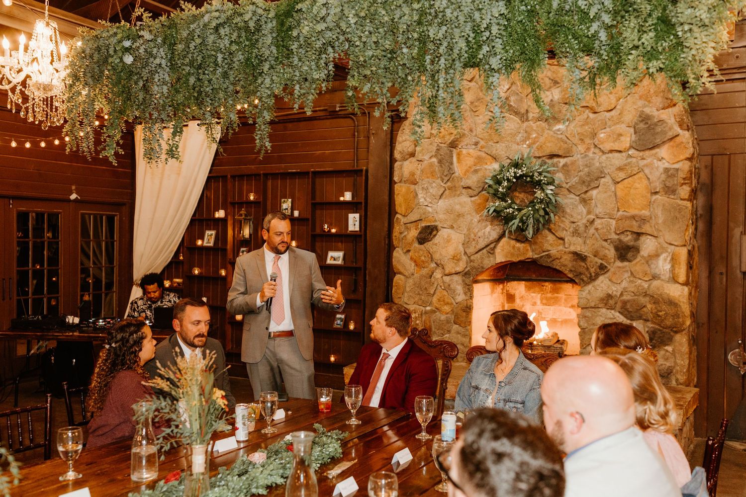 Man in a gray suit gives a toast to seated wedding guests at a rustic reception with a stone fireplace and greenery garland.
