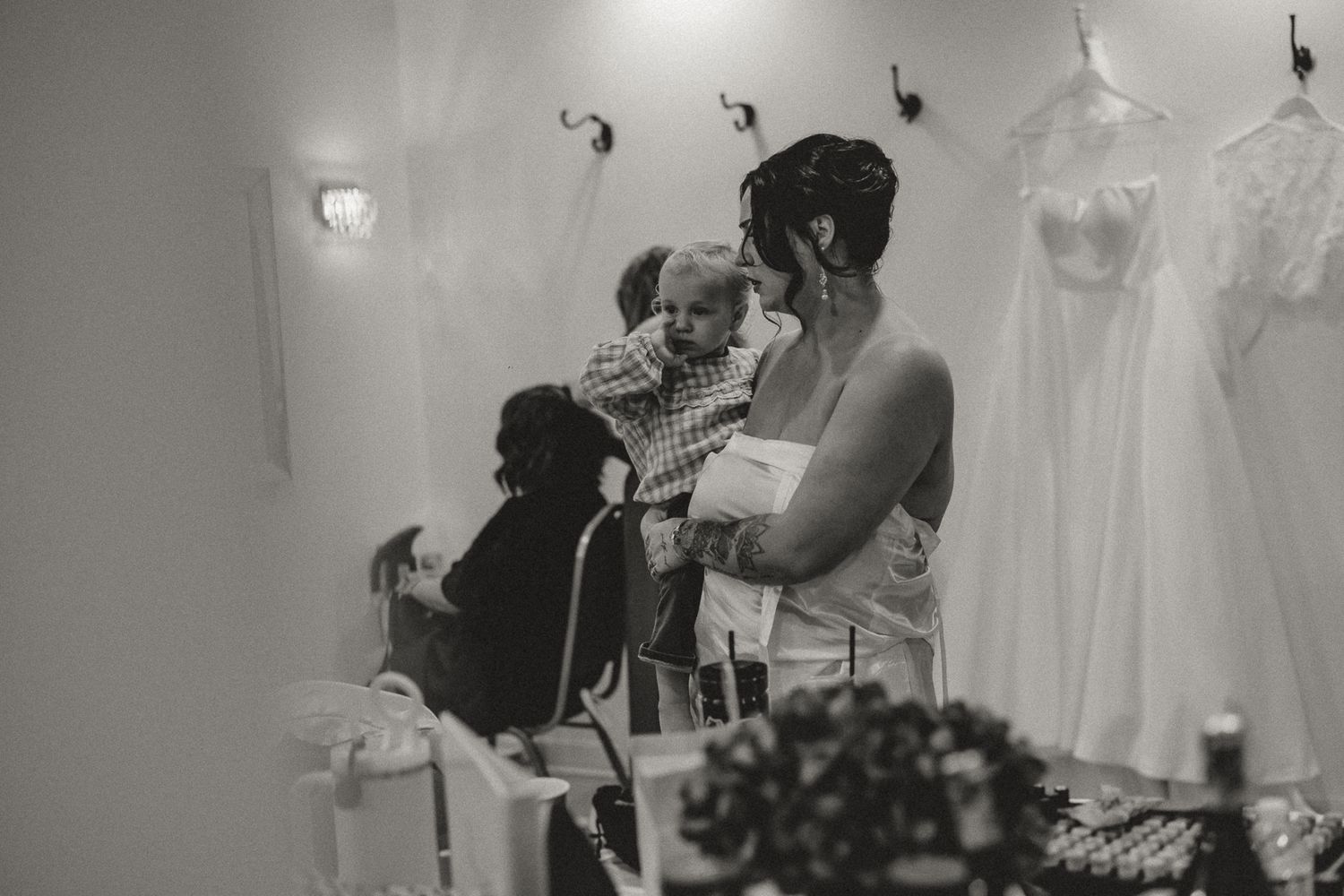 Black and white photo of a woman holding a baby in a bridal boutique with wedding gowns hanging in the background.