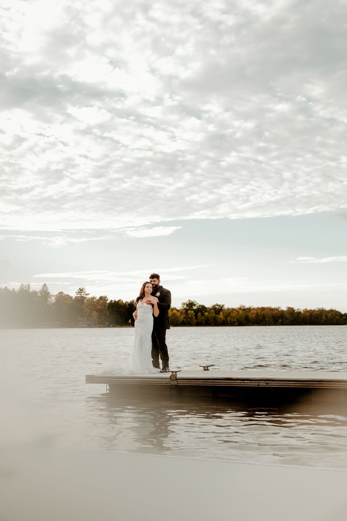 Bride in a white gown and groom in a dark suit embrace on a wooden dock overlooking a calm lake beneath a soft, cloudy sky.