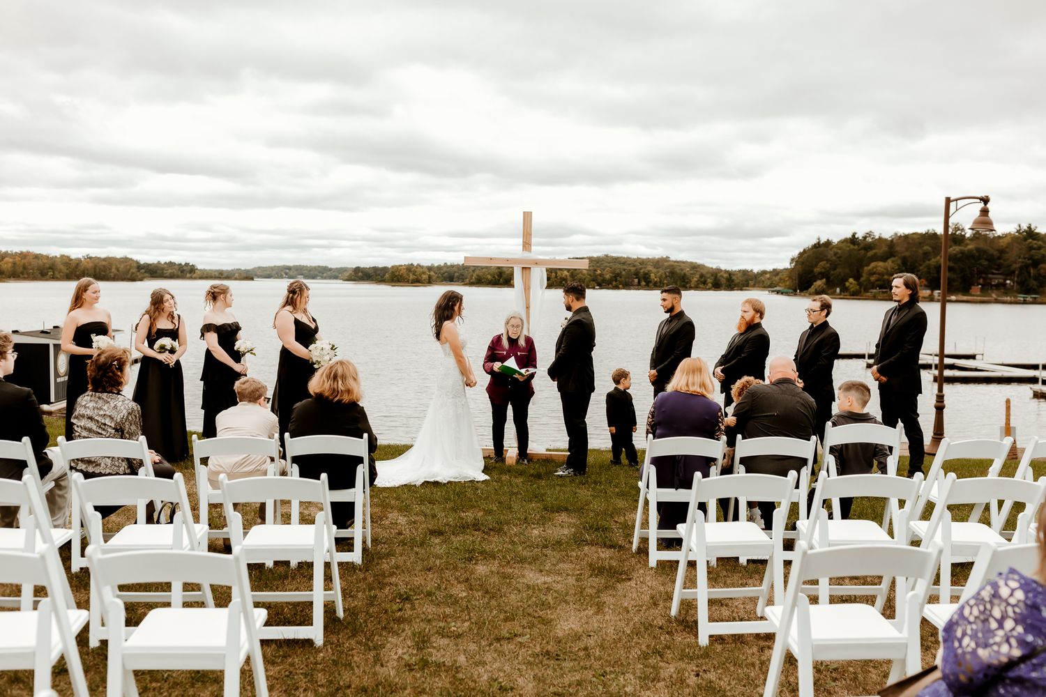 Bride, groom and officiant stand before a wooden cross during an outdoor lakeside wedding ceremony as guests sit in white chairs.