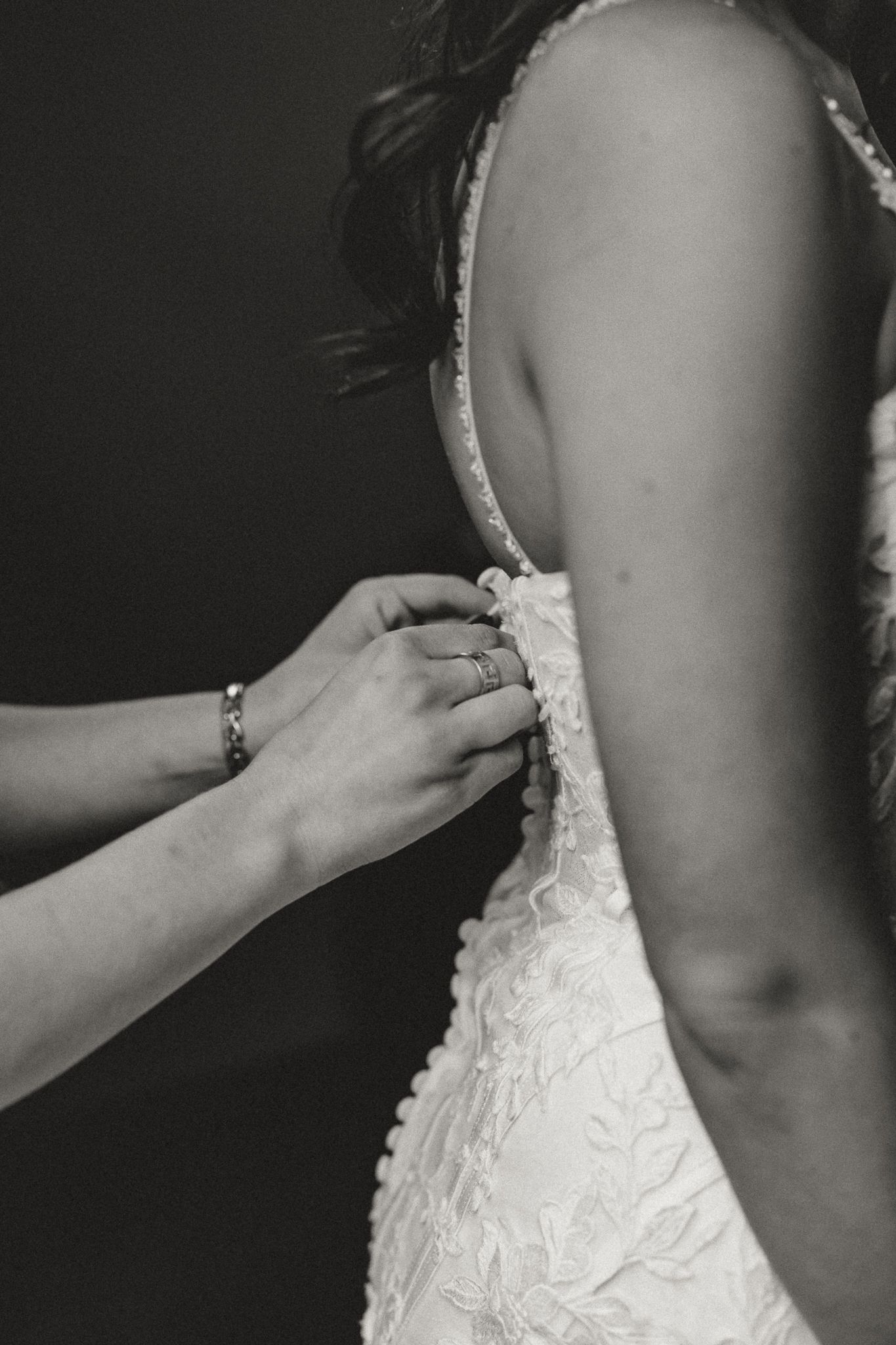 Black and white photo of a bride in a lace wedding gown as a bridesmaid fastens the buttons on the back of her dress.