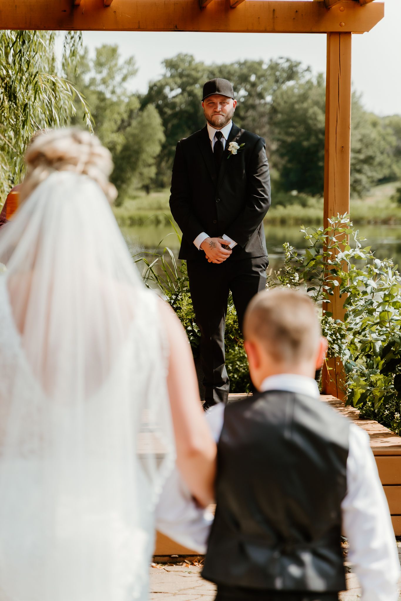 Groom watches bride walk down aisle at outdoor wedding ceremony under wooden pergola near pond.