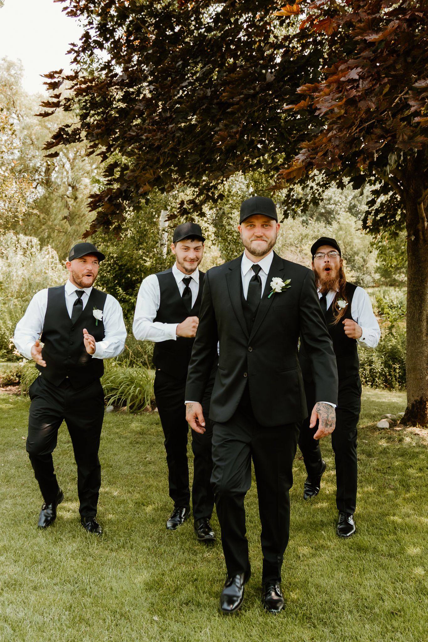 Groom in black suit with boutonniere leads groomsmen in black vests and caps walking outdoors on green lawn.