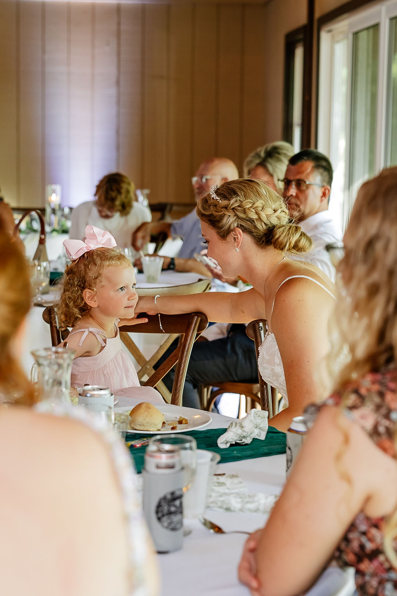 A woman in a wedding reception leans down to interact with a young girl wearing a pink bow at a decorated dining table.