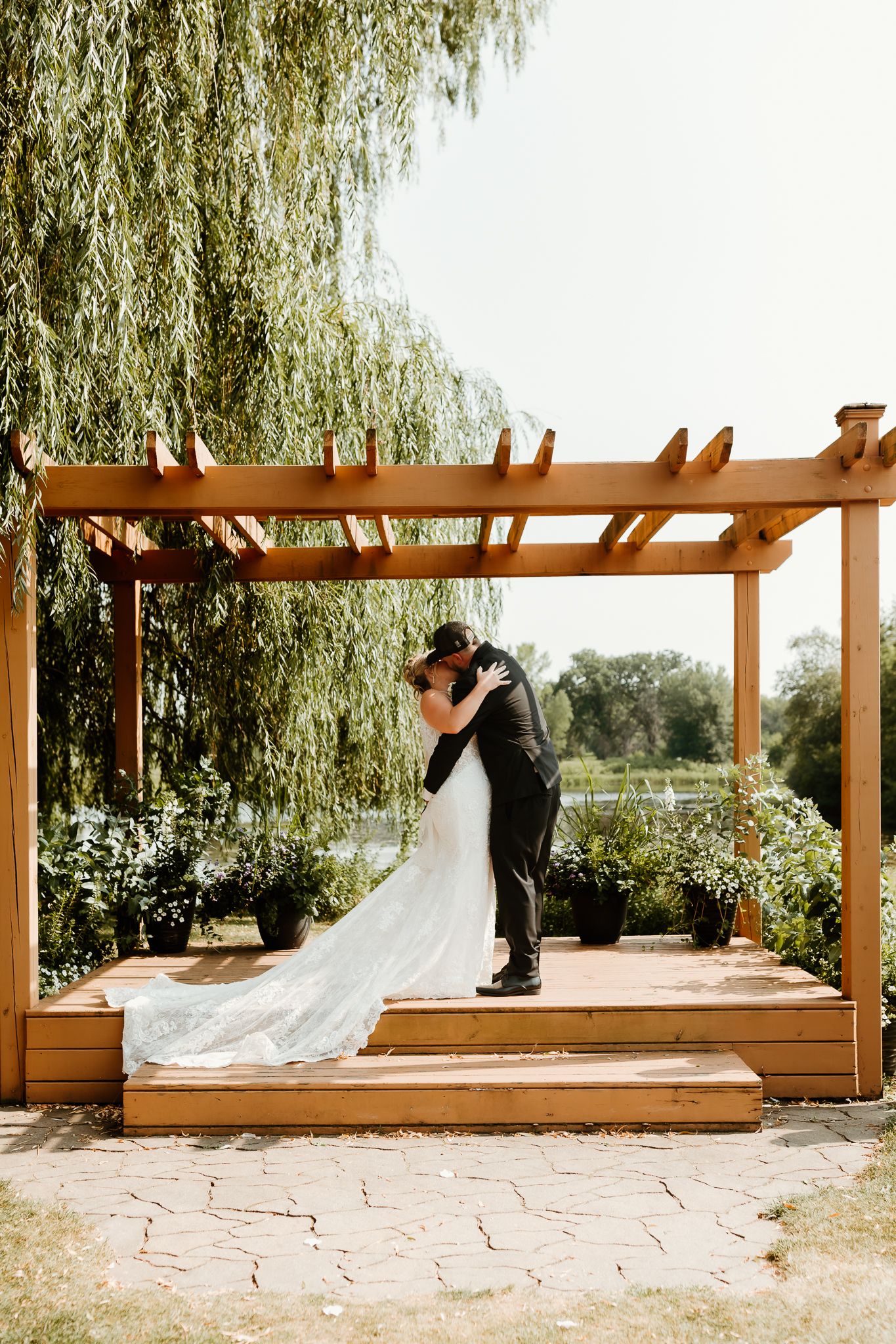 Bride and groom share a kiss under a wooden pergola surrounded by lush greenery on their wedding day.