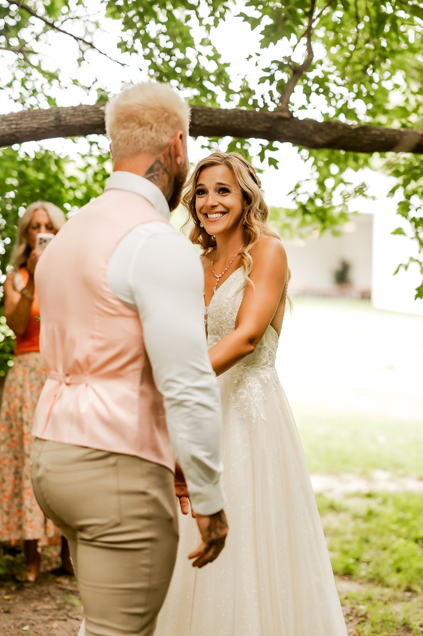 Bride in white lace gown smiles joyfully at groom in peach vest during outdoor wedding ceremony.