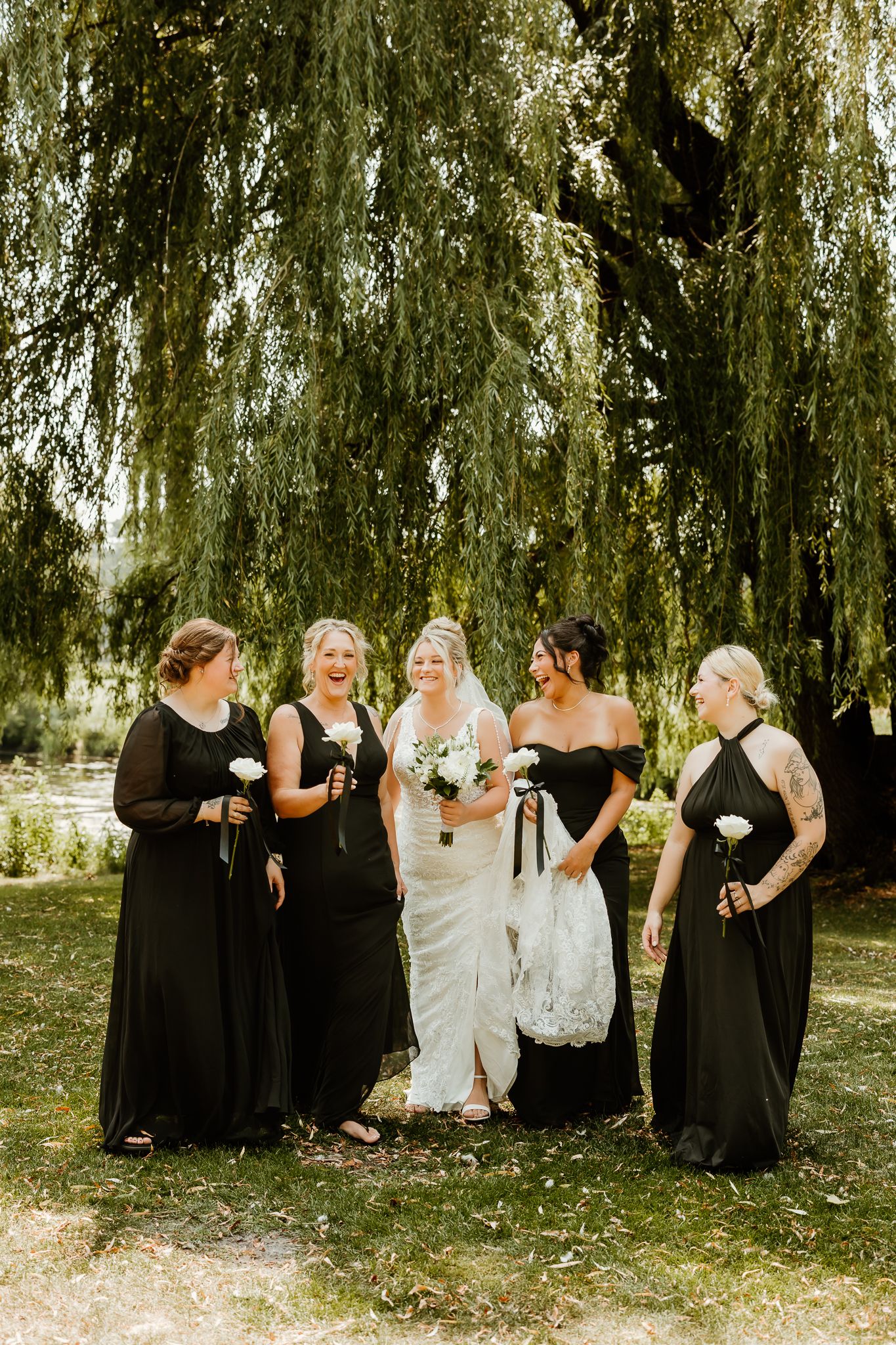 Bride in white lace gown with four bridesmaids in black dresses under a weeping willow tree outdoors.