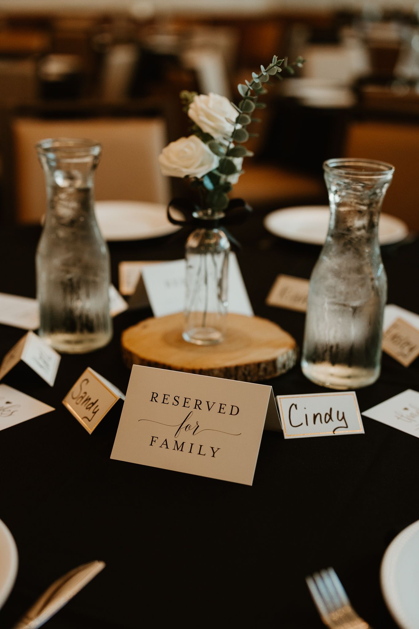 Elegant wedding reception table with Reserved for Family sign, glass vases, white roses, and name cards on black tablecloth.