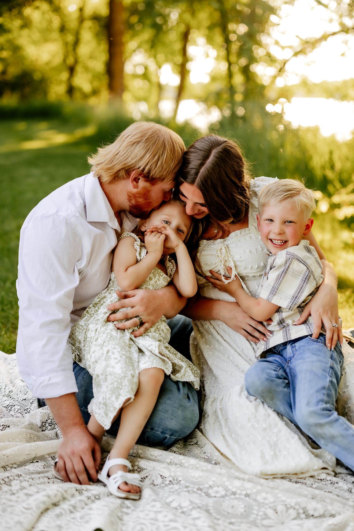 Family of four sharing joyful hugs and kisses on a blanket in warm golden sunlight outdoors.